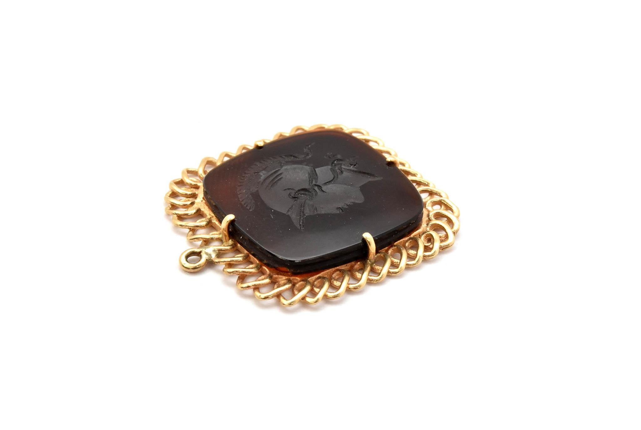 This pendant features a brown stone carved with an intaglio. The intaglio is set into a 14k yellow gold pendant setting. The piece measures 26x29mm, and it weighs 5.0 grams.