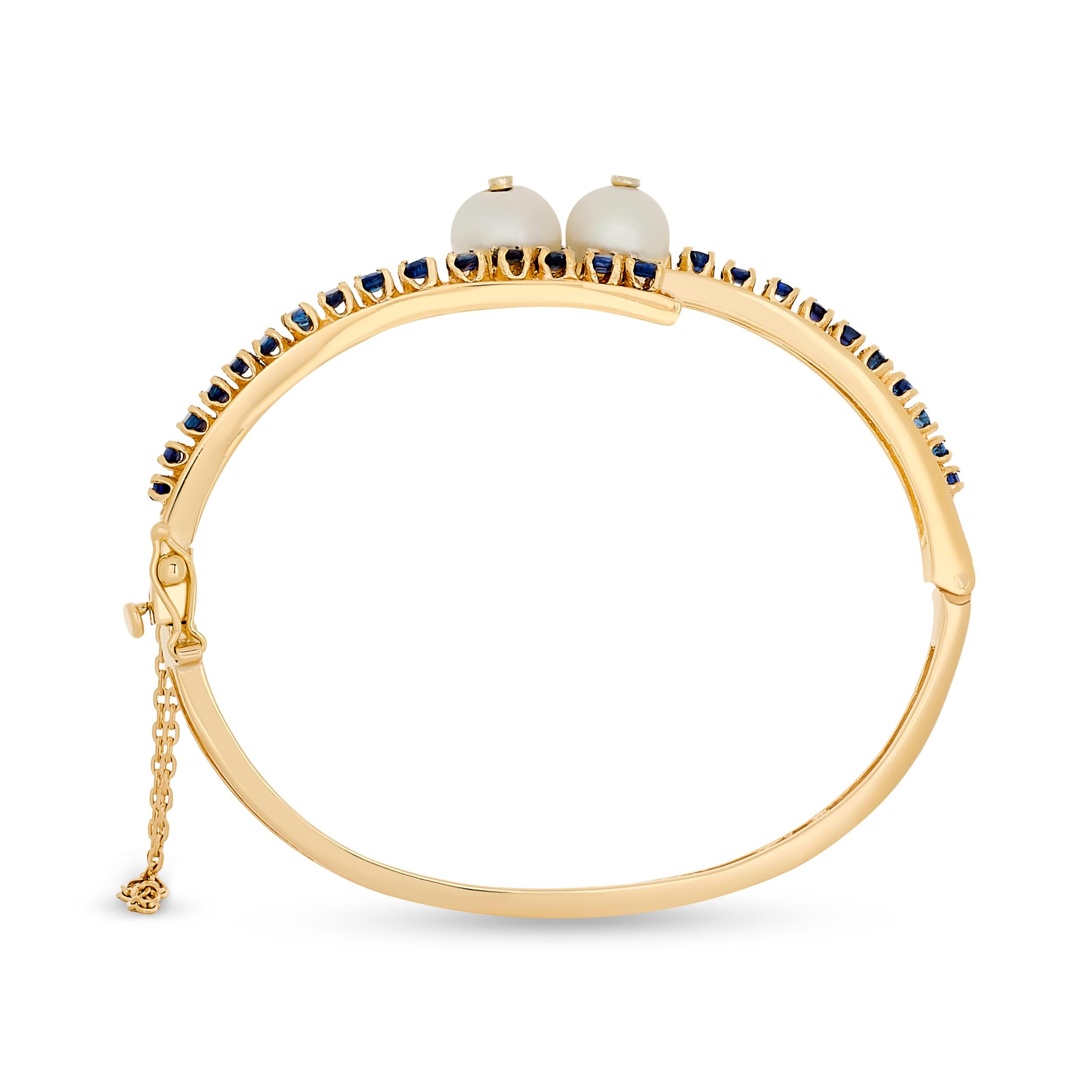 A contemporary 14k yellow gold bangle featuring two luminous pearls nestled between parallel rows of vibrant blue sapphires, creating a striking and elegant design.

6.5