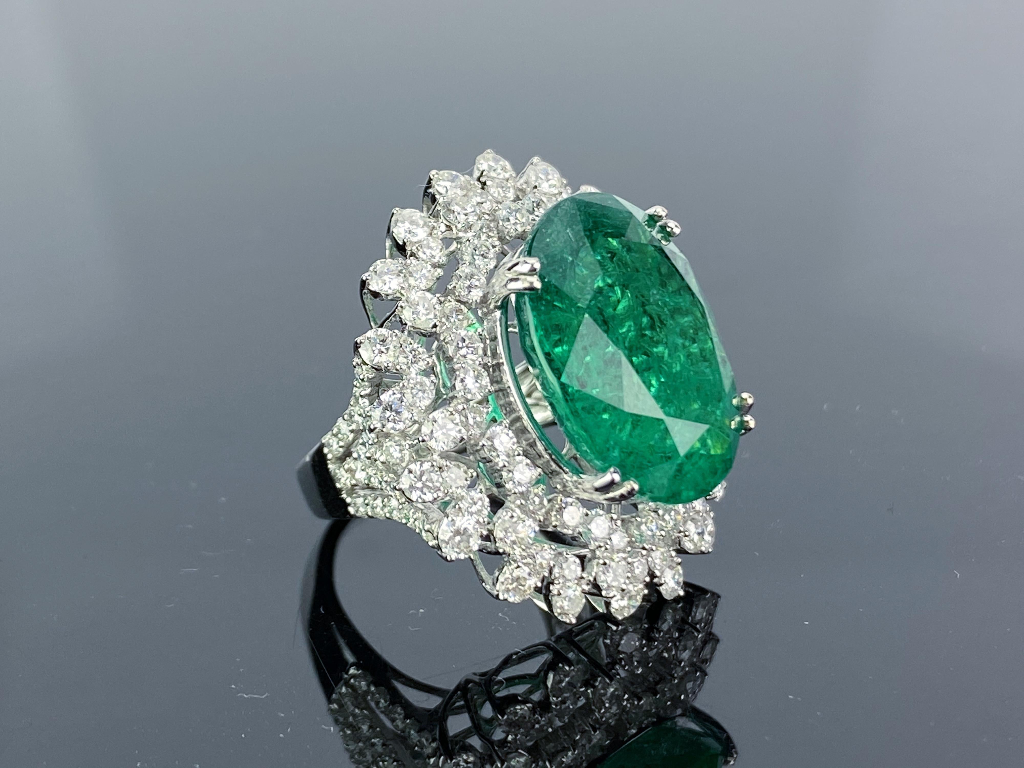 An eye-catching cocktail ring, with a 14.03 carat transparent natural Zambian Emerald centre stone sorrounded with diamonds. The emerald is transparent with good lustre and colour, with few inclusions. All set in 18K white gold. Currently a ring