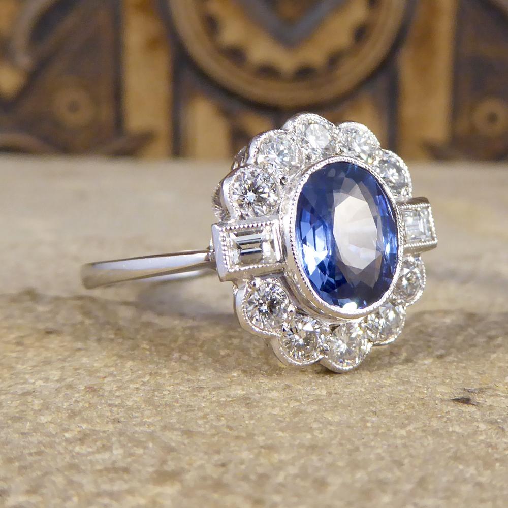 This beautiful blue oval cut Sapphire weighs 1.40ct and is held in a rub over collar setting with a millegrain detail. The cluster of Diamonds consist of five round brilliant cut Diamonds over the top of the Sapphire and five below, separated by two