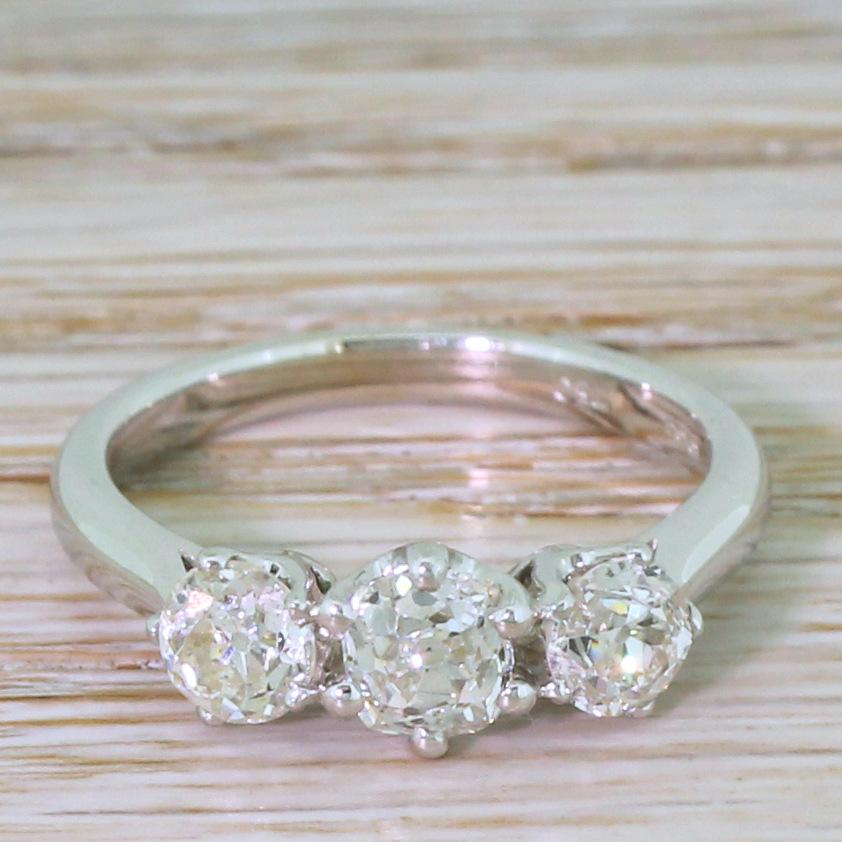 Three fiery and bright diamonds make up this old cut trilogy ring. A slightly larger cushion shaped old cut in the centre is flanked by a pair of round old cuts, all set in classic coronet collets leading to a tapering D-shaped white gold