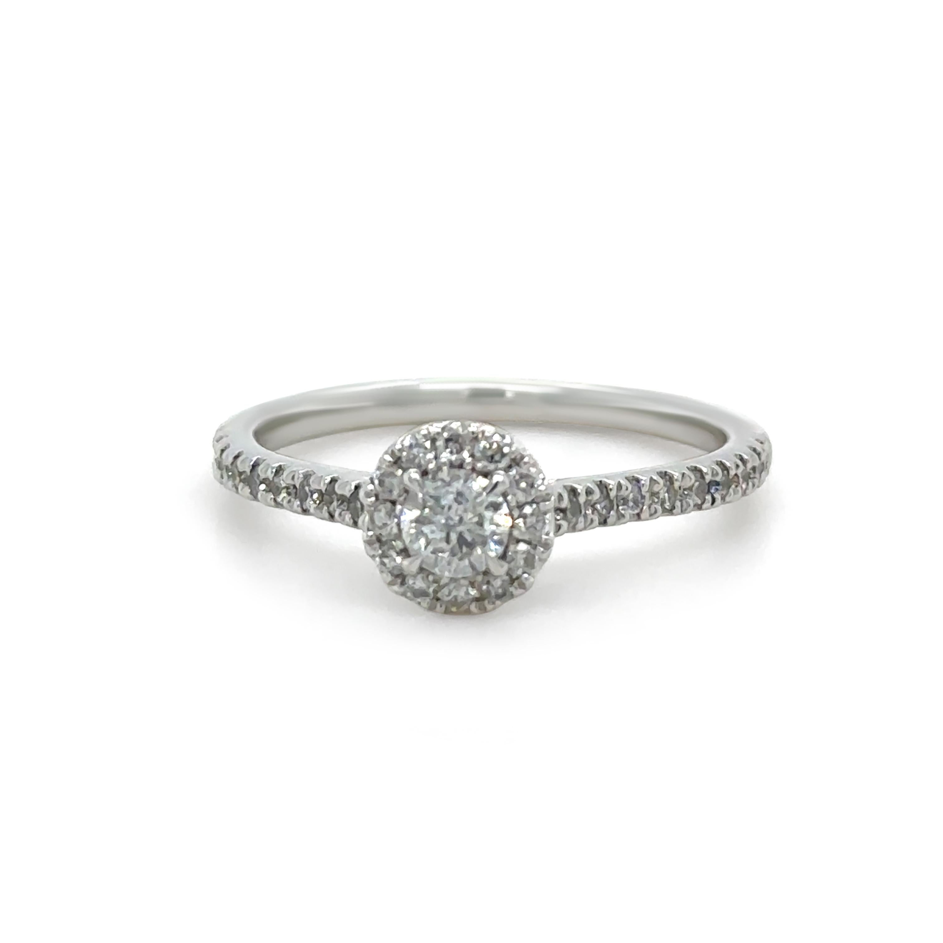 Cette bague de mariage en or blanc 14ct est ornée d'un diamant central rond de taille brillant de 0,23ct, serti de quatre griffes et entouré d'un halo de diamants ronds sertis de griffes. Le poids total combiné des diamants n'est pas inférieur à