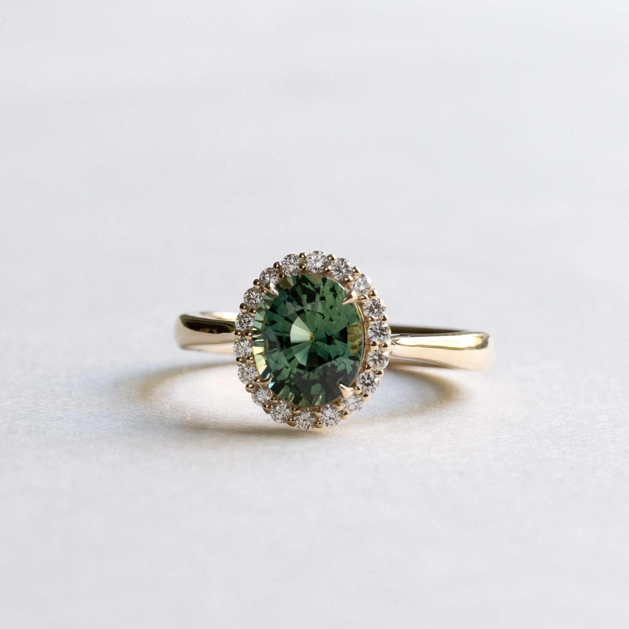 Green sapphire adorned with diamond halo. 

Metal: 14K Yellow Gold
Stone: Green Parti Sapphire
Stone Weight: 2 Carat 
Stone Shape: Oval 
Stone Size: 6mm x 8mm 
Accent Stone: Round Diamond
Diamond Color and Clarity: G+, VS
Stone Size: 1.5mm
Total