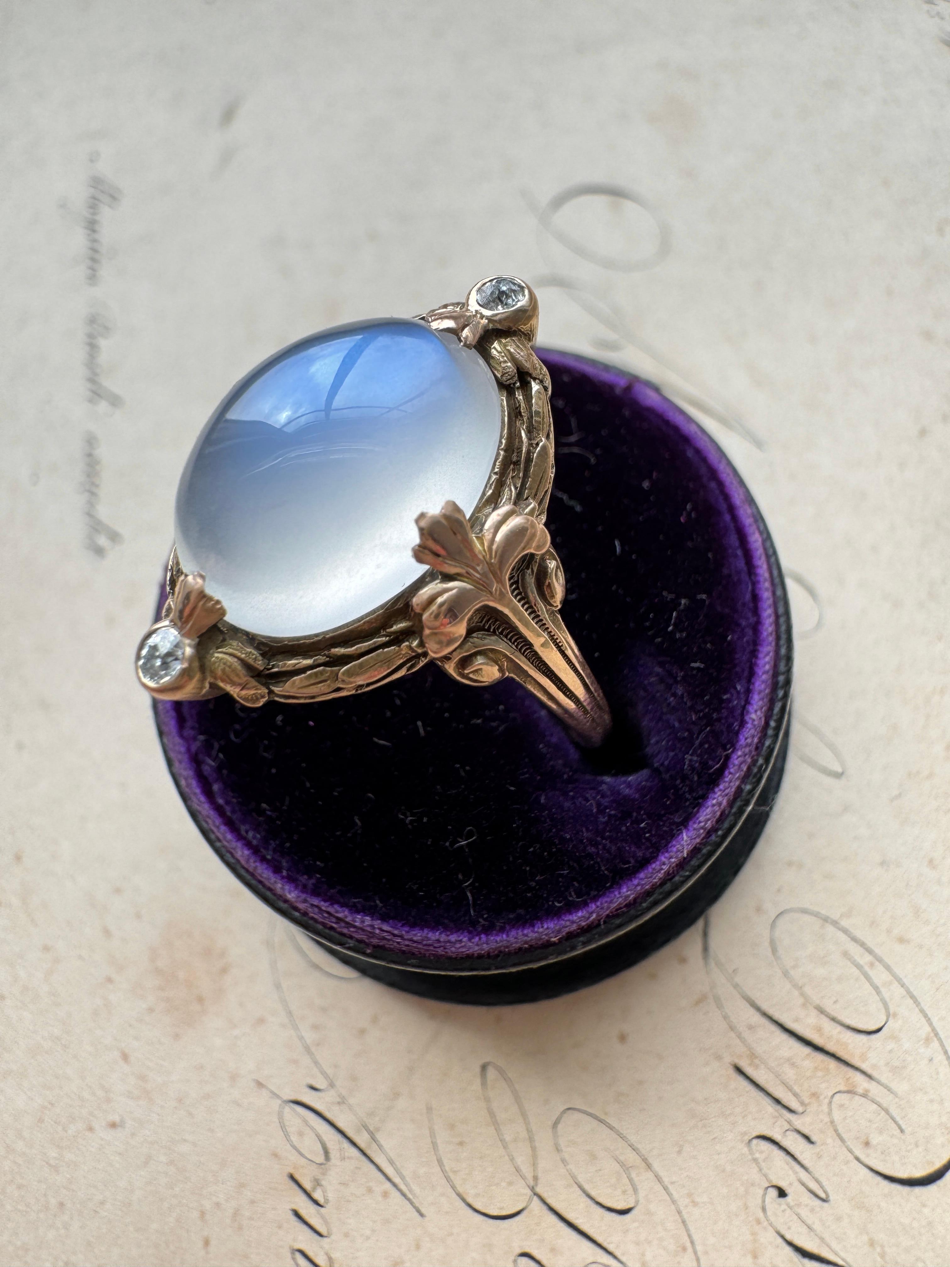 Dating from the early 20th century, this enchanting Art Nouveau ring showcases a luminous blue-flash moonstone elegantly framed by a sculpted laurel leaf border with fleur-de-lis shoulders and two twinkling diamonds. Currently a ring size 7