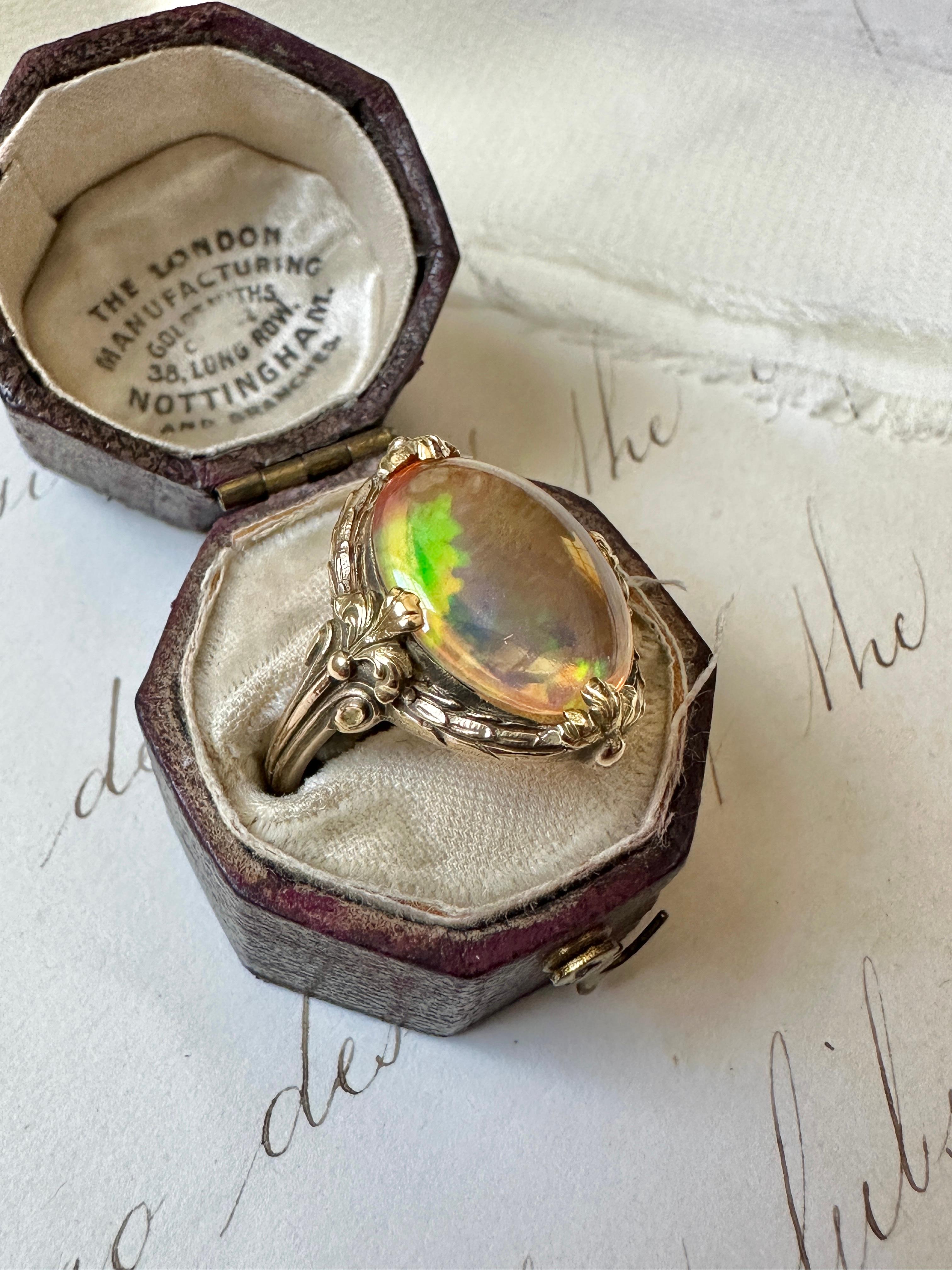 Dating from the early 20th century, this delightful Art Nouveau ring centers on a glowing opal filled with confetti like flashes of bright yellow, vibrant red, brilliant blue and neon green. The opal is presented in a sculpted laurel leaf border