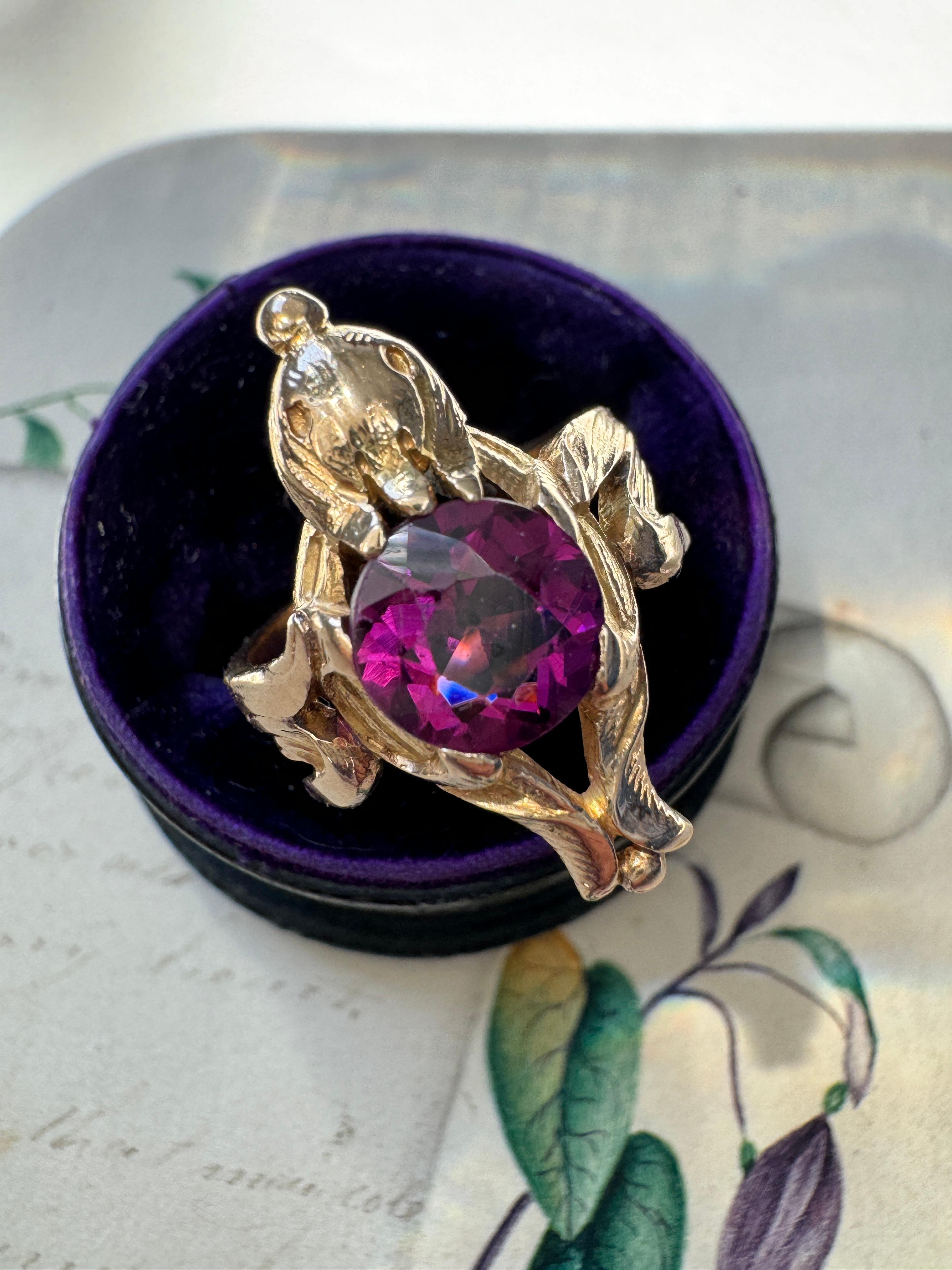 This charming Art Nouveau bird ring centers on a glowing 1.55 carat pinkish-purple rhodolite garnet held by a pair of sinuous leaves that double as stylized wings. Circa 1900, crafted in burnished 14k gold, hand textured throughout. Currently a ring