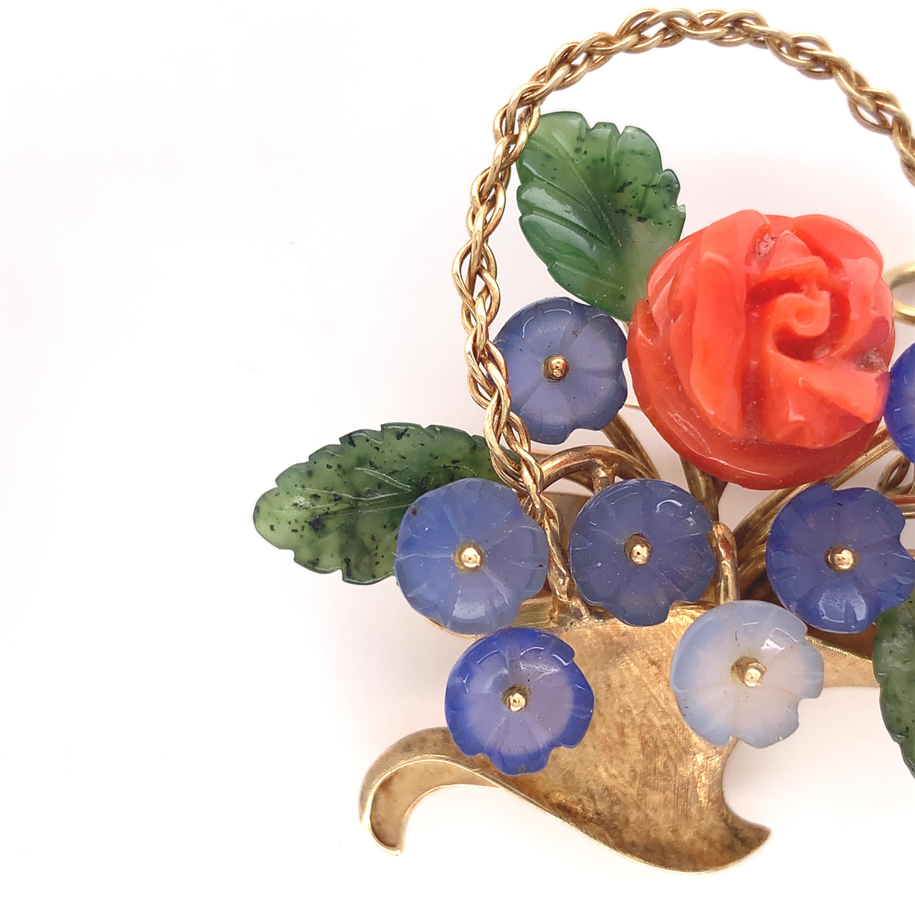 14K yellow gold pin featuring a basket of carved flowers with leaves. There are 10 carved bluish-grey chalcedony flowers, one large carved coral rose and 3 jade leaves. The pin is handmade, and the handle is braided gold wire. The carved chalcedony