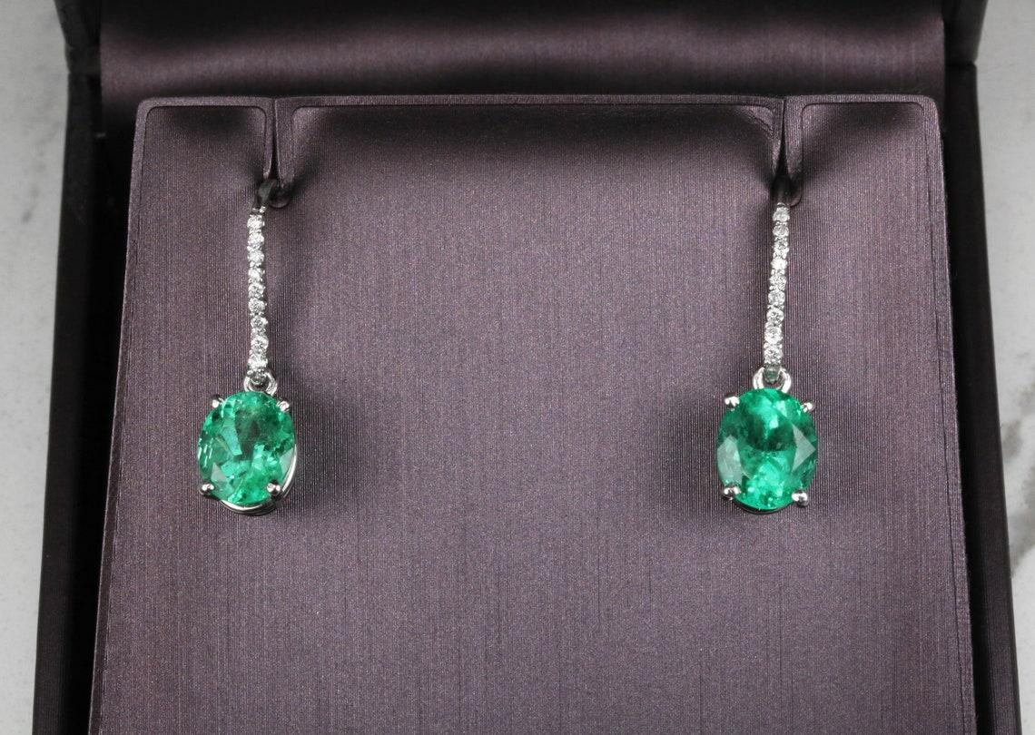 An immaculate pair of HIGH grade and quality genuine Colombian oval emeralds and white diamond drop/dangle gold earrings in solid gold. Dexterously Handcrafted in solid 14K white gold, these earrings feature two vivacious fine quality natural
