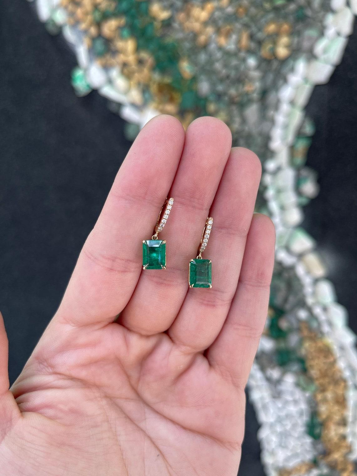 Introducing our exquisite Emerald Dangle Earrings, a stunning display of elegance and sophistication. Two mesmerizing emerald cut emeralds, totaling over four carats in combined weight, dangle gracefully from knife-edge huggie U-shaped hoops adorned