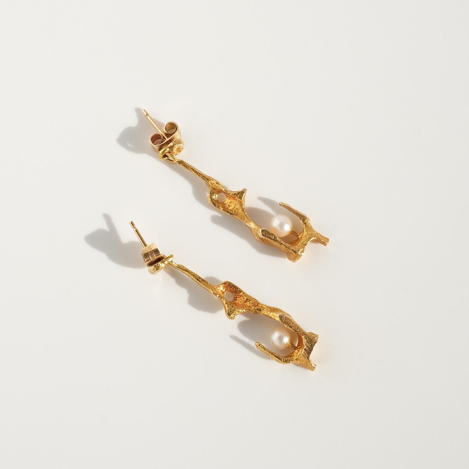 These 14 karat gold dangling earrings, known as At the source (Sw. Vid källan) feature an organic, sculptural design with a brushed finish that creates a soft, matte effect. Each earring has a delicate cultured pearl nestled within the lower part of