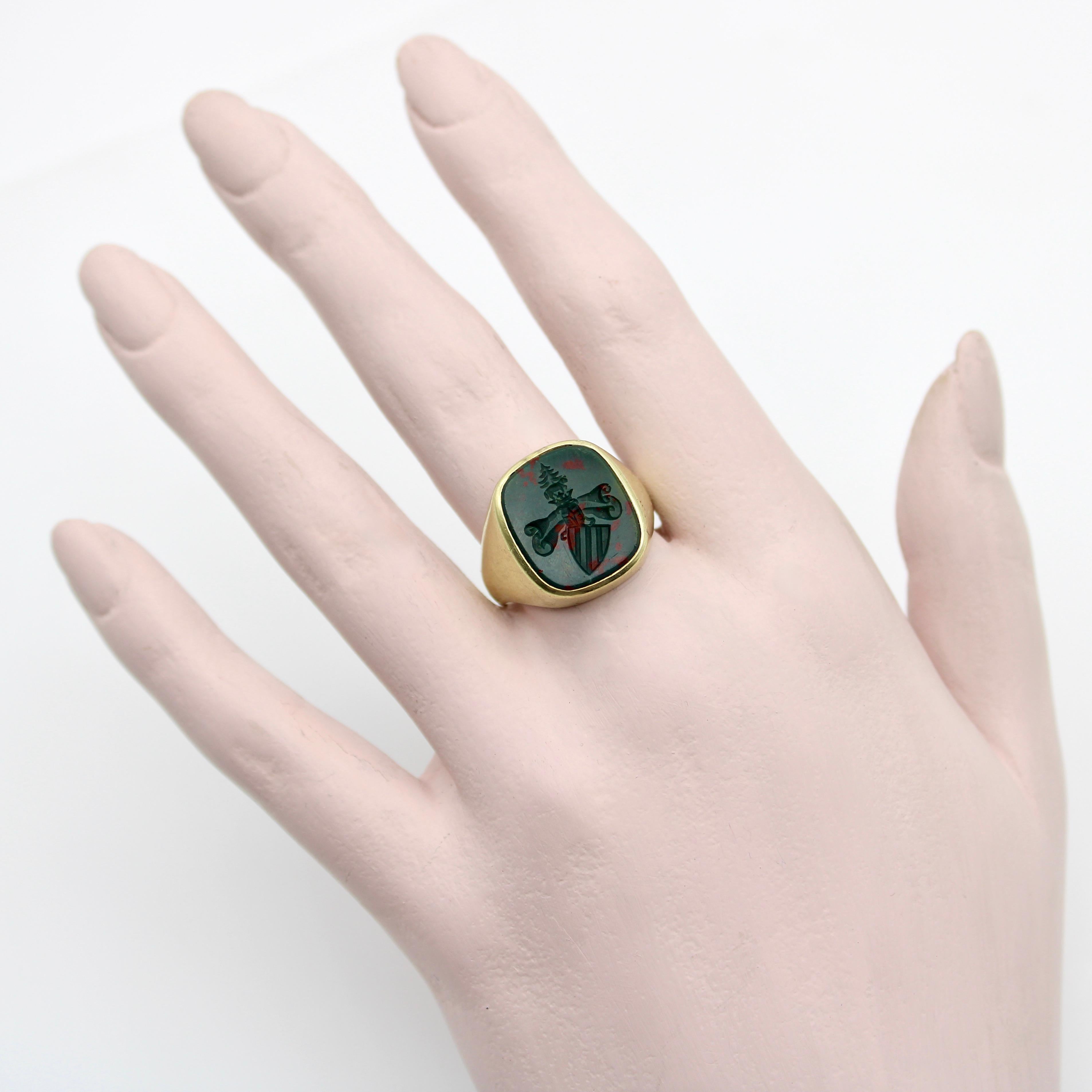 This 14k gold Edwardian intaglio signet ring contains an esoteric image—a unique family crest with mysterious imagery. Carved into bloodstone, a striped shield sits on a diagonal, with a scroll looming above it. It is topped with a crown, a pine