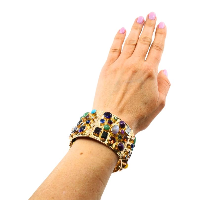 A 14k gold bracelet, featuring cabochon cut gemstones.
The bracelet has a distinctive look, made up of multiple hinged sections. The gold has a warm rosy tone, complementing the array of cabochon cut gemstones scattered across the surface. The