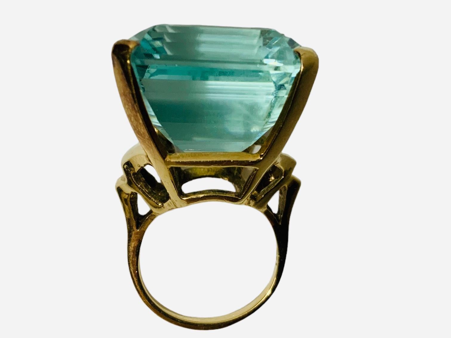This is a 14K yellow gold Emerald Cut Aquamarine cocktail ring. The blue greenish Beryl Aquamarine is faceted and set in four gold prongs. Its measurements are 25.51 x 21.71 x 16.26 mm and It weighs 61.23 carats. The side walls between the prongs