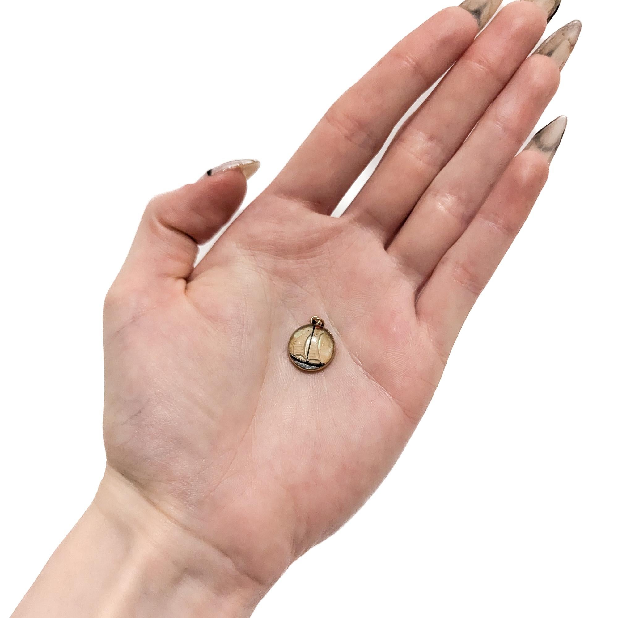 Un charme en or antique et en cristal d'Essex.

En or jaune 14 carats.

Au centre, un cabochon en cristal d'Essex décoré d'un voilier de style sloop arborant un drapeau rouge sur fond de nacre.

La partie supérieure est munie d'un anneau de sécurité