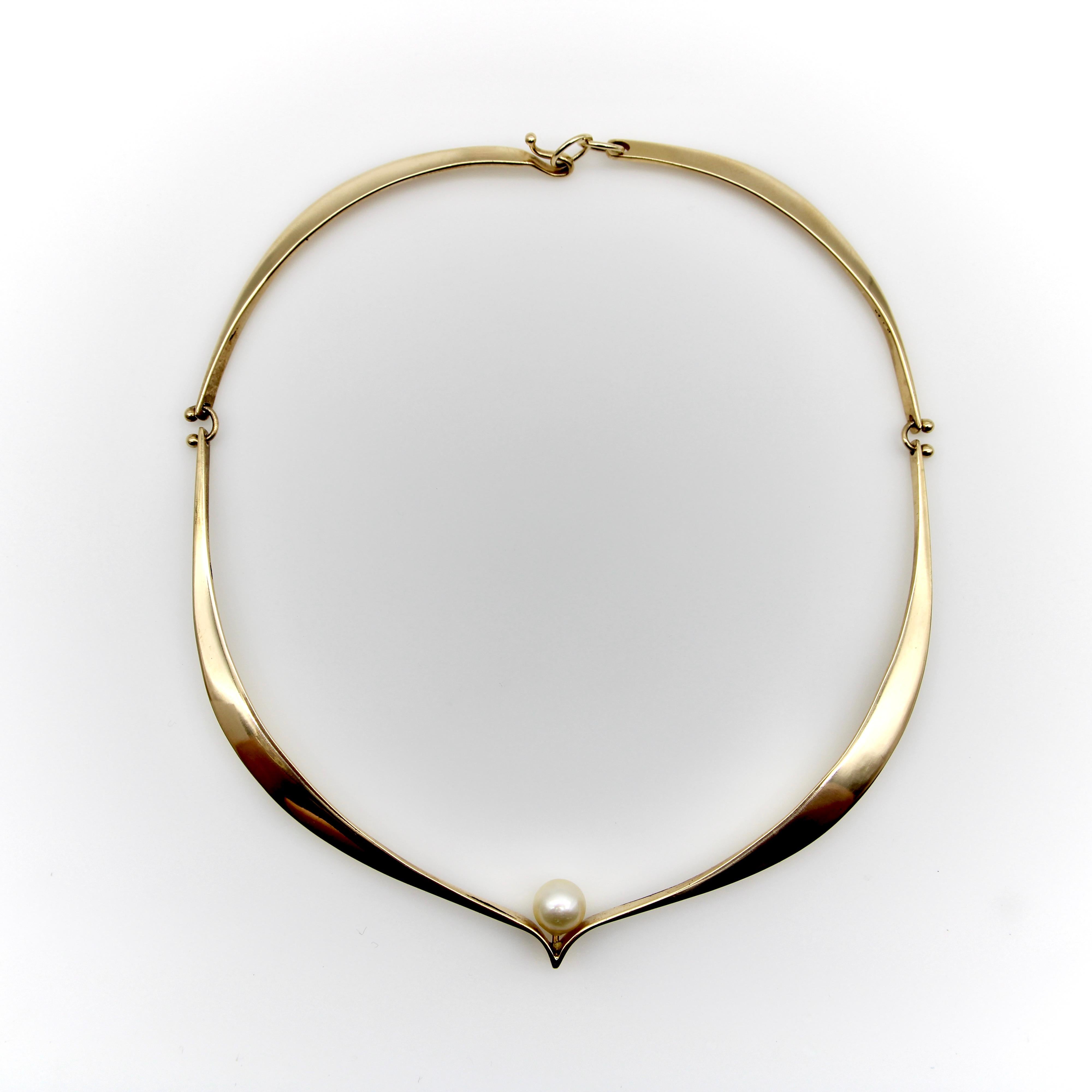 This 14k gold signed Ed Wiener necklace has a sculptural form that graces the collarbone in an elegant wave. At the center of the necklace, a pearl is nestled into a small chevron that places it perfectly along the clavicle. It is hinged with tiny