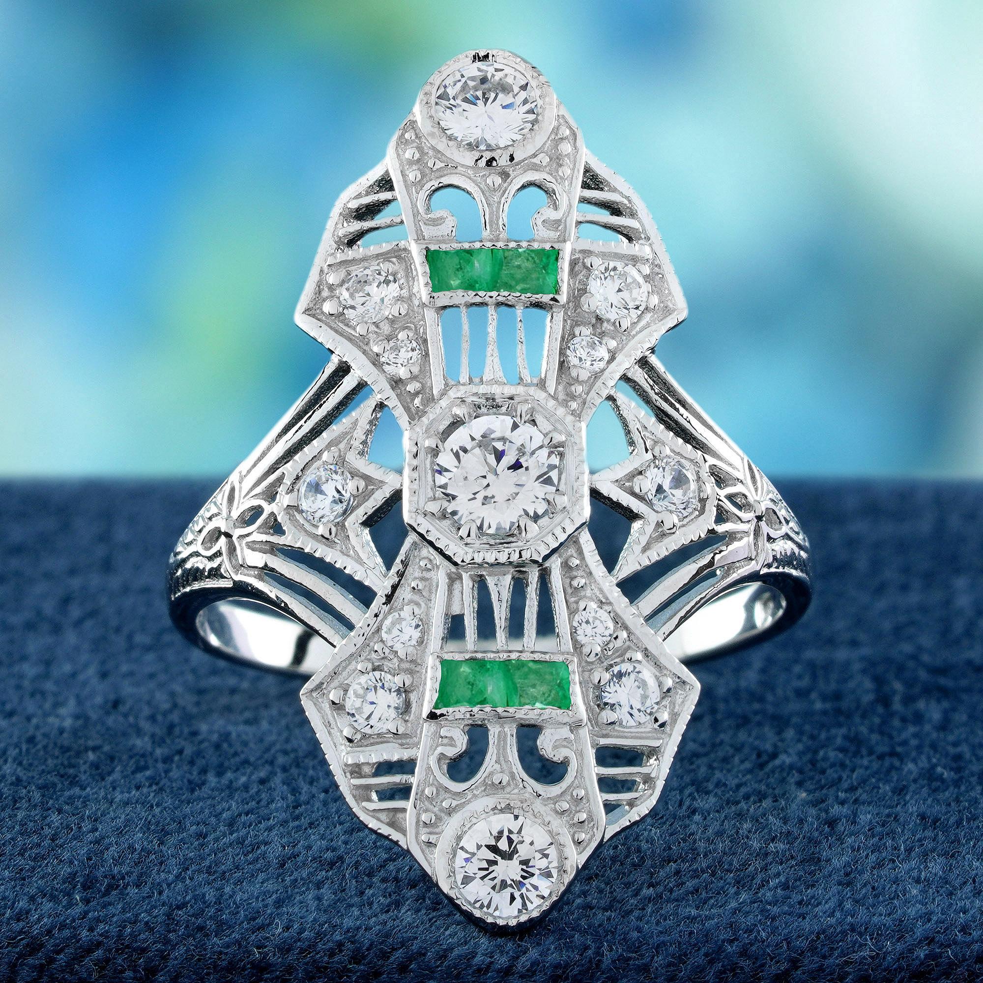 A brilliant round diamond sits at the center, radiating timeless sparkle and sophistication. French-cut emeralds accent the top and bottom of the setting, adding a striking pop of rich green and vintage flair. The intricate vintage-inspired