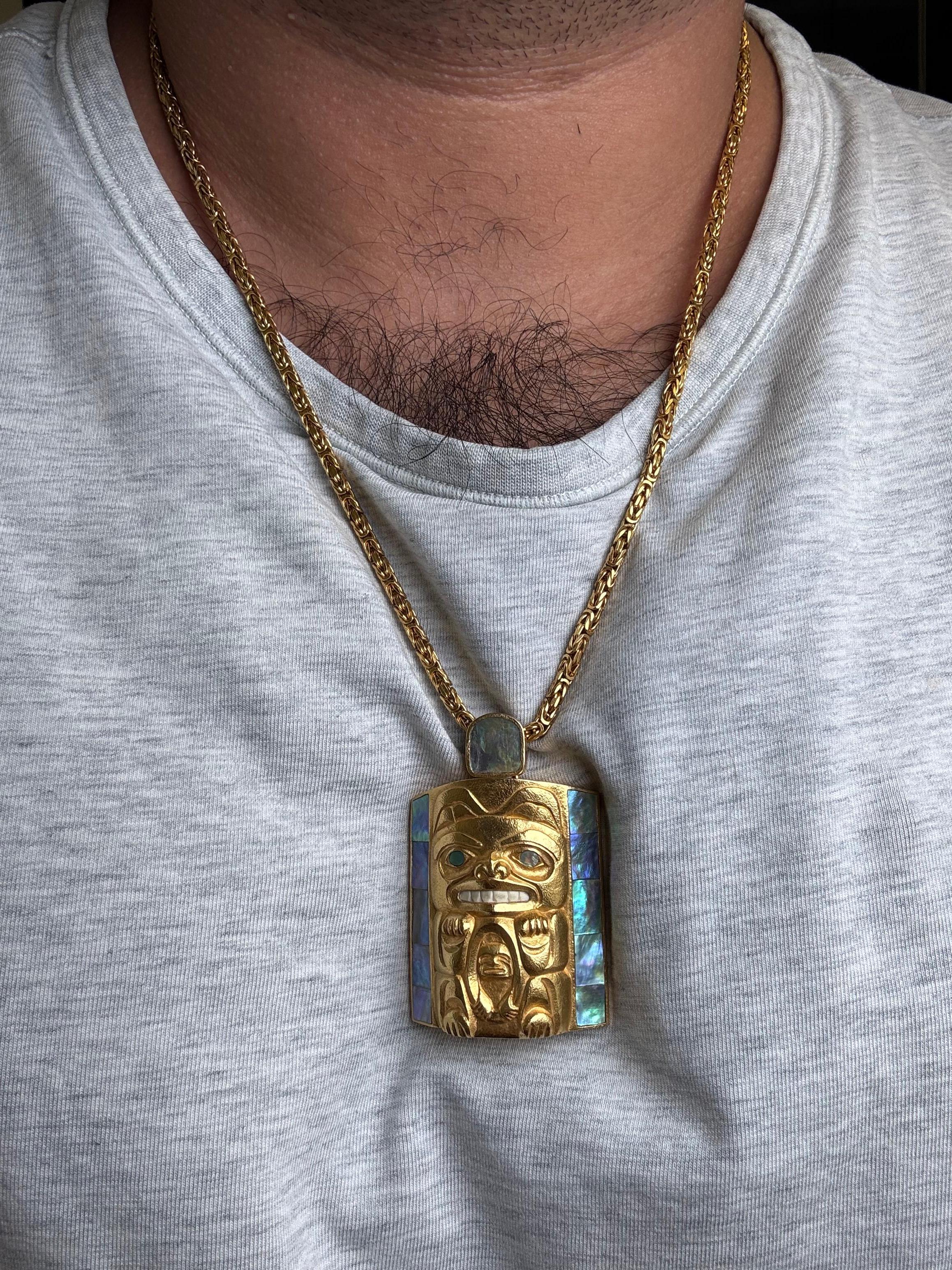 A remarkable handcrafted pendant inspired by Northwest Coast Indigenous art, depicting a stylized totem figure rendered in richly textured 14-karat yellow gold. The design is framed by vivid abalone shell inlays, which display iridescent blue,