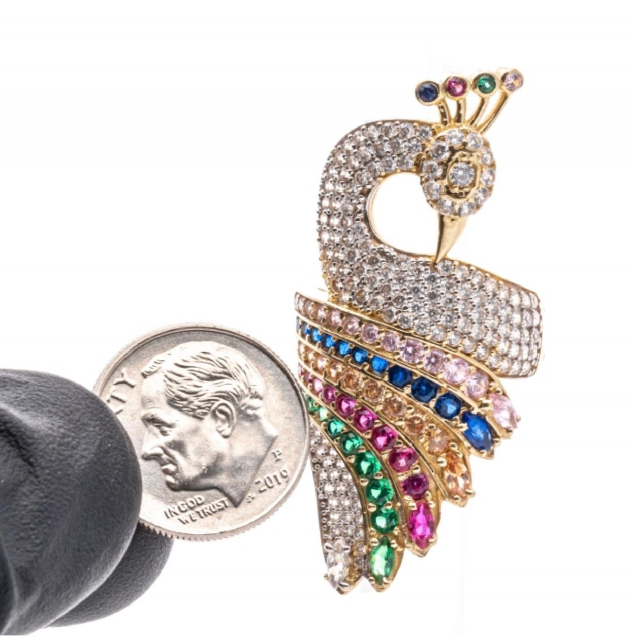 This majestic 14k yellow gold ring is a figural representation of a peacock, with a flat profile, and pave set throughout with pave set, round brilliant cut CZ stones. In the body of the ring is a sweep of color, featuring rows of pale pink, royal