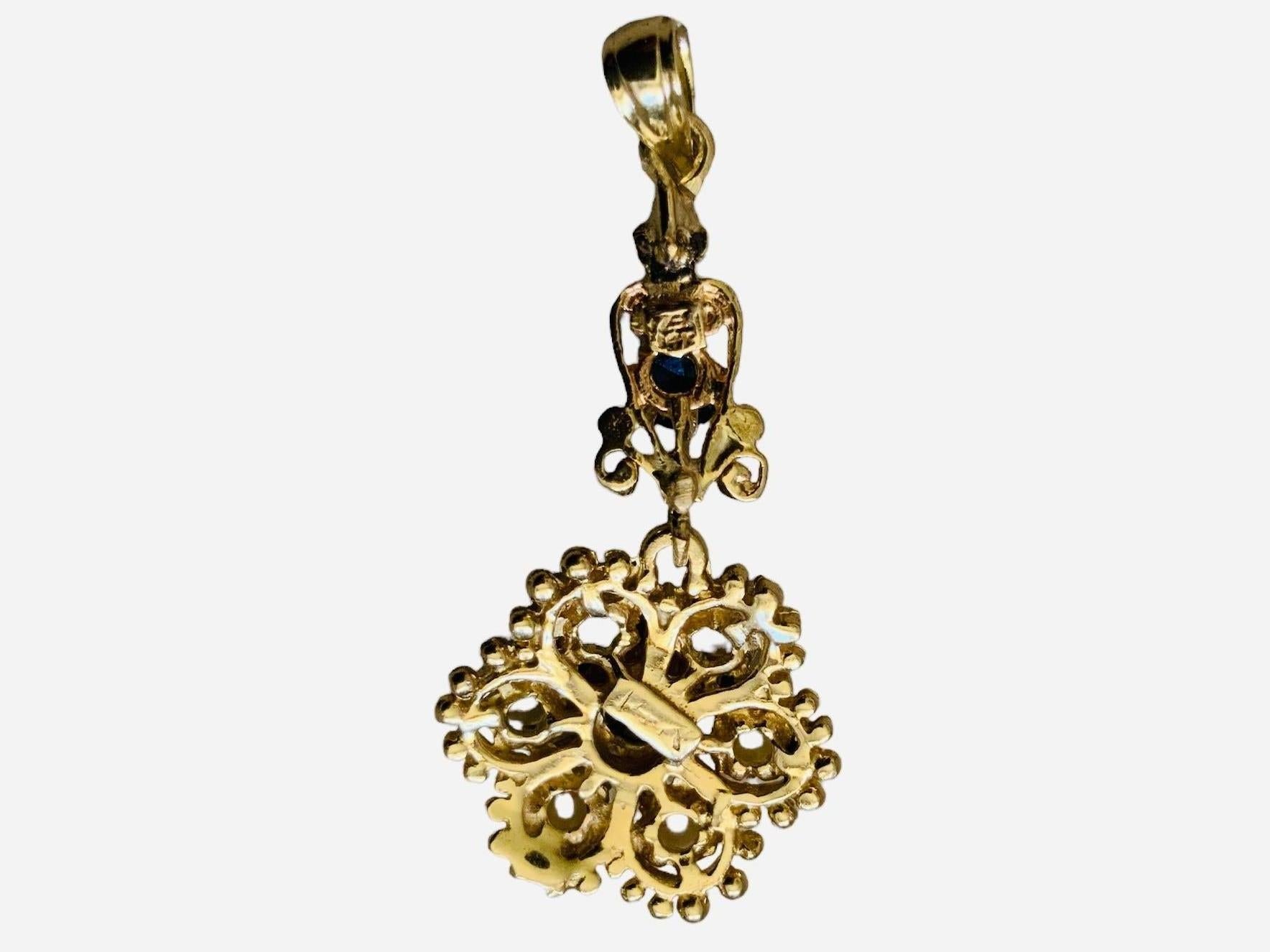 This is a 14K gold Pearl and Dark Blue Topaz pendant. It depicts a gold flower pendant adorned with six pearls in prong setting and a dark blue topaz also in prong setting in the center. Another smaller dark blue topaz is above the flower.