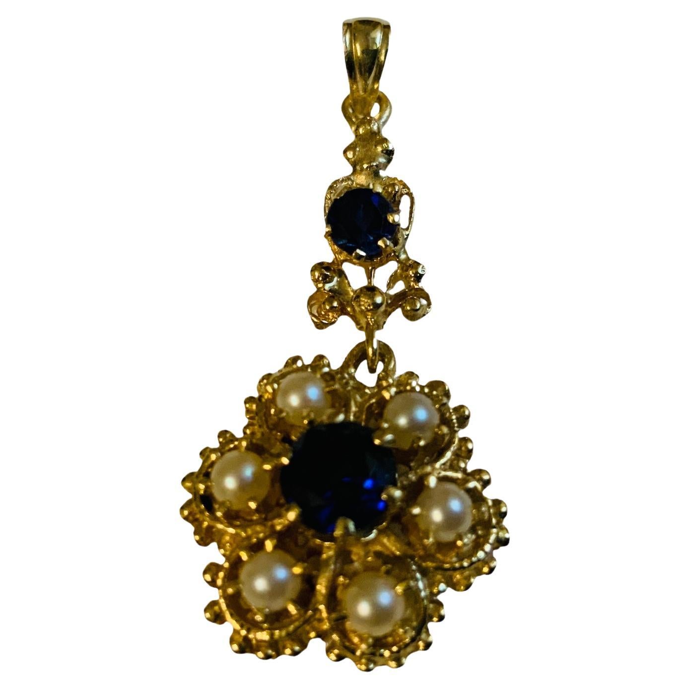 This is a 14K gold Pearl and Dark Blue Topaz pendant. It depicts a gold flower pendant adorned with six pearls in prong setting and a dark blue topaz also in prong setting in the center. Another smaller dark blue topaz is above the flower.