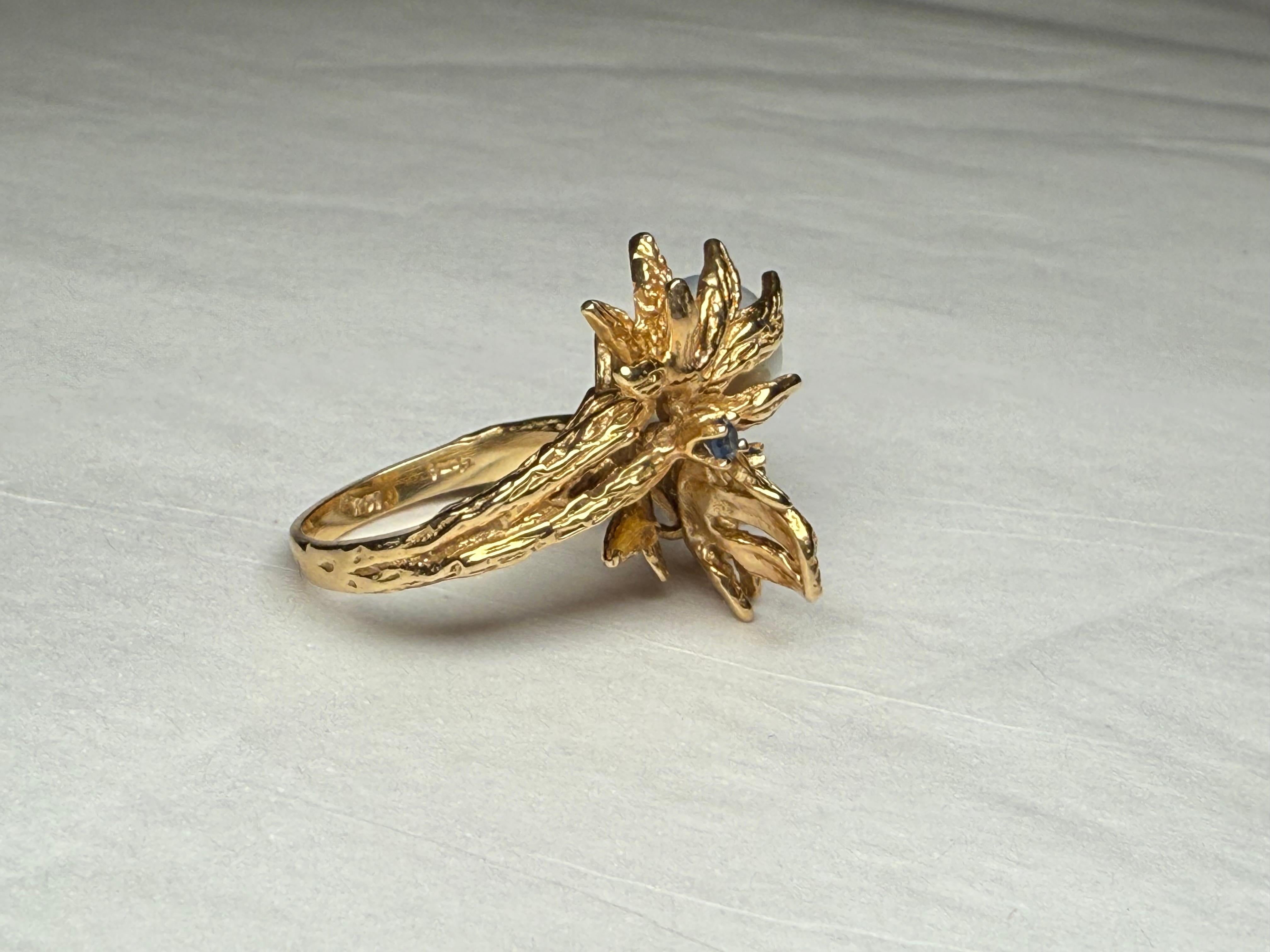 This is a 14K Gold Pearl and Sapphires Crossover Ring. It depicts a wrap around ring ending with two flowers with several petals holding a blue gray pearl and each one of them enhanced with a sapphire in a prong setting. The ring is hallmarked 14K