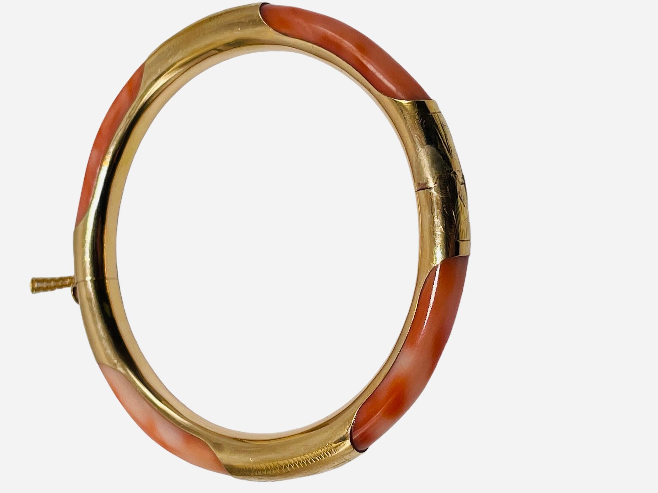 This is a 14K gold Coral hinged bangle/bracelet. It depicts a Pink Coral bangle adorned with bands of gold engraved with some branches of leaves. The bracelet has a cylindrical box closure with a heart clasp and safety chain. It is hallmarked 585