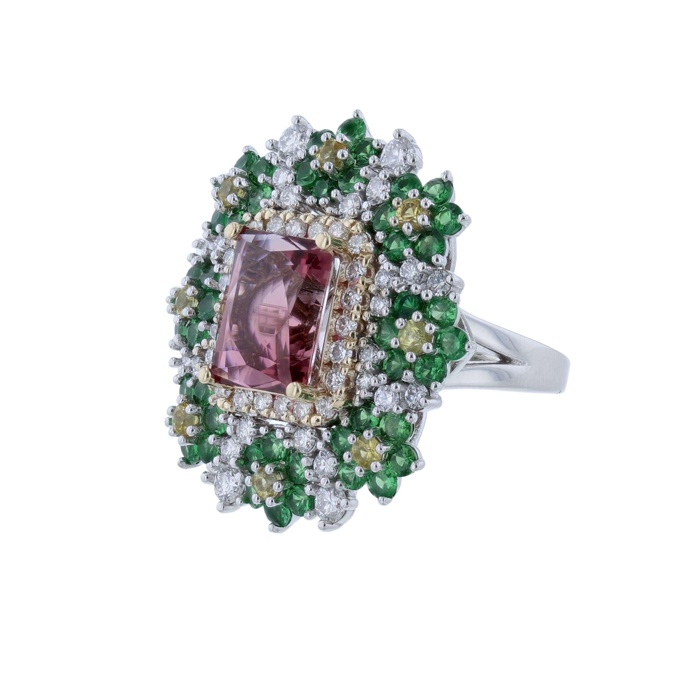 This ring is made of 14K white and yellow gold. It features 1 emerald cut pink tourmaline center stone weighing 5.40. Surrounded by 50 round cut diamonds weighing 1.45 carats, 48 round tsavorite weighing 2.67 carats, and 8 yellow sapphires weighing