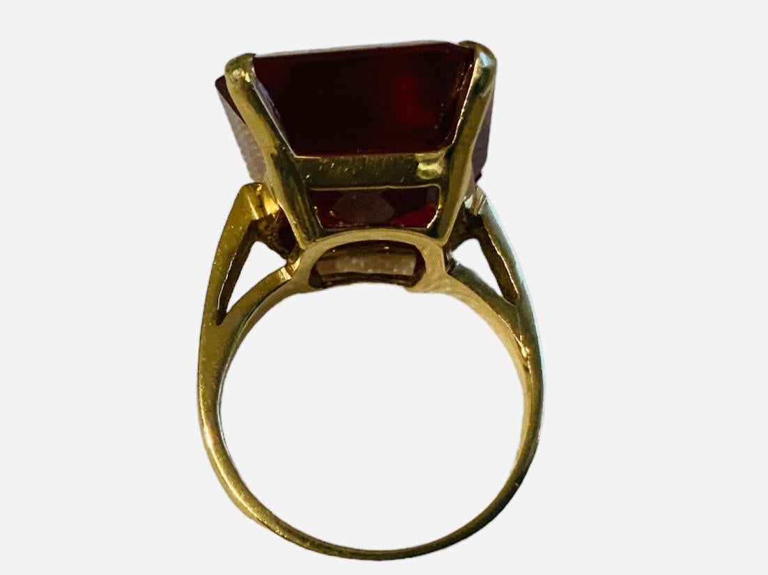 This is a 14K yellow gold Emerald Cut Ruby cocktail ring. The ruby is faceted and set in four gold prongs. Its measurements are 0.75in x 0.5in. The side walls between the prongs are pierced in a rectangular shape. It is signed 14K inside one of the