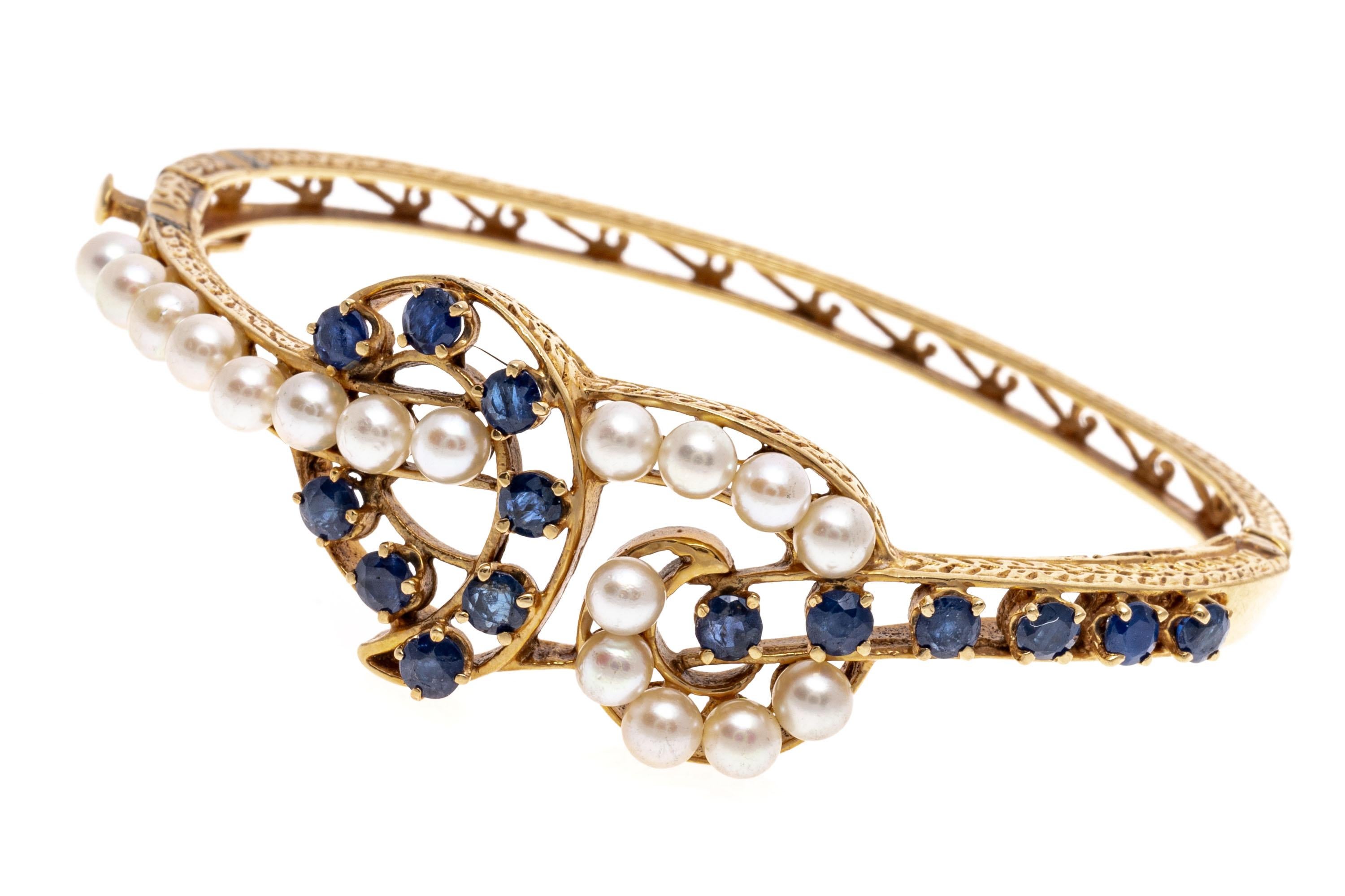 A graceful 14K yellow gold bangle bracelet with intertwining bands of bright white cultured pearls and indigo blue sapphires. The back of the band is accented is a delicate open filigree like design. Box style closure with safety latch for added