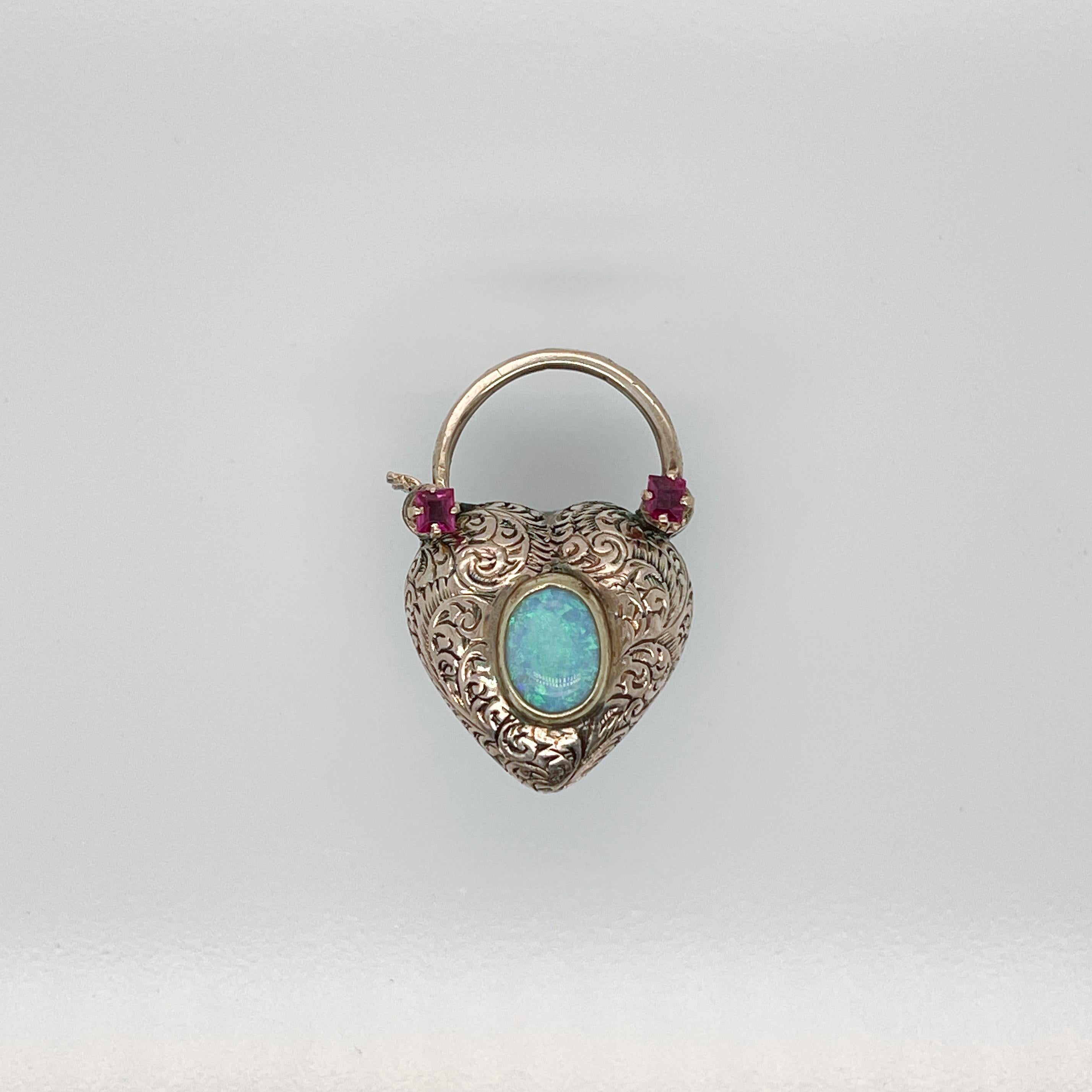 A very fine 14 karat gold, turquoise, opal, and garnet charm.

In the form of a puffy heart-shaped lock with a hinged, curved catch to the top.

One side is bezel set with a heart shaped turquoise cabochon and the reverse is both bezel set with an