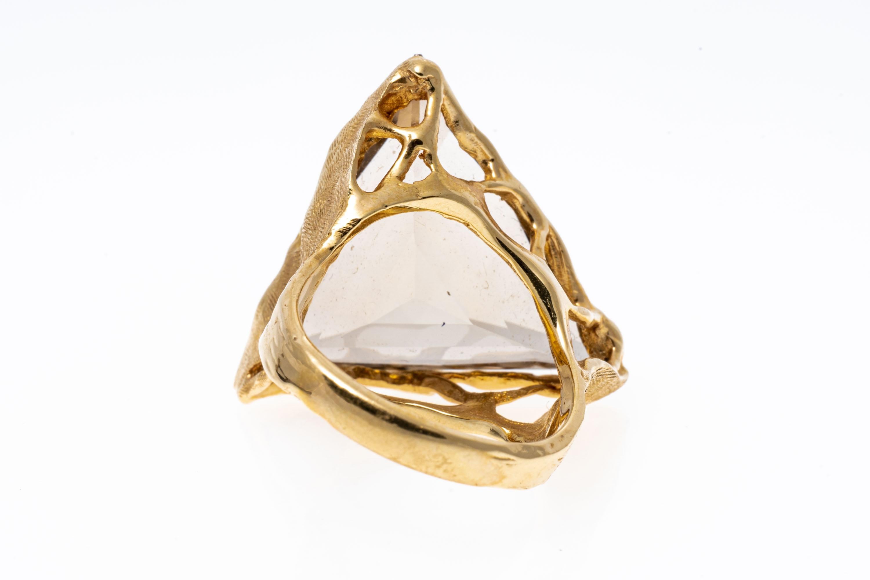 14k yellow gold ring. This contemporary ring features a striking large trillion faceted, pale brown smoky quartz ring, approximately 19.36 CTS, set into a free form style mounting with large chased foliate themed shoulders.
Marks: 14k
Dimensions: