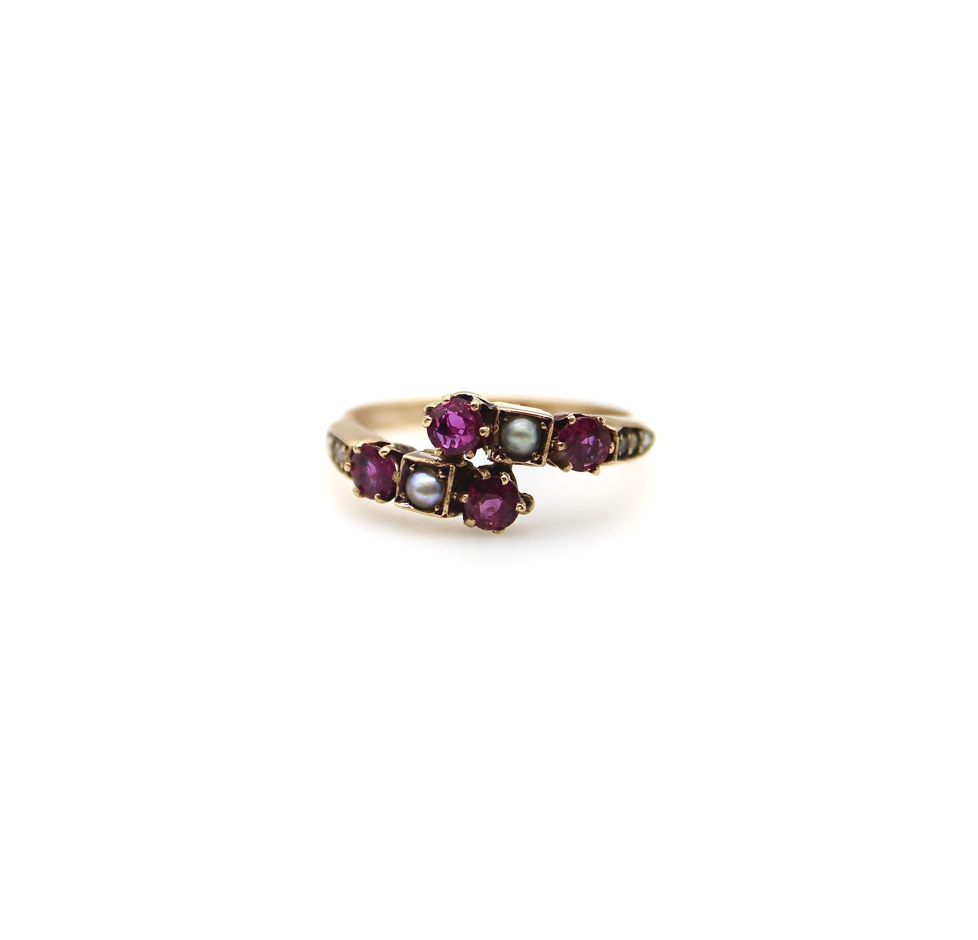 A lovely Victorian ring with classic motifs that are feminine and delicate. This 14k gold band mirrors itself and meets at the center in a cluster of gemstones—a milky white seed pearl flanked by two rubies. The gemstones are prong set, with the