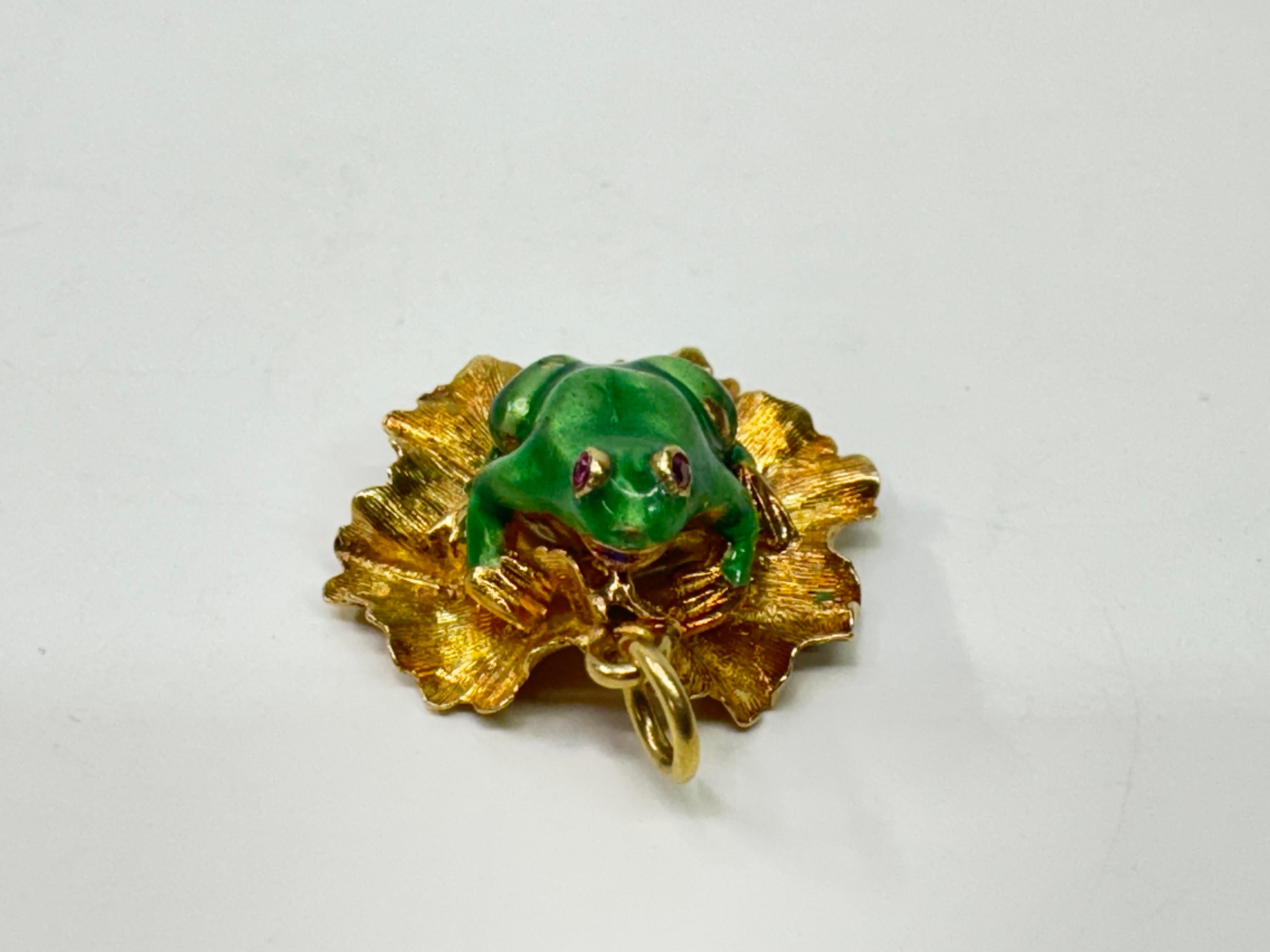 Questo affascinante ciondolo a forma di rana in oro 14k è caratterizzato da un vibrante smalto verde, che siede con grazia su una ninfea in oro giallo. Gli occhi della rana sono ornati da scintillanti gemme di rubino, che aggiungono un tocco di