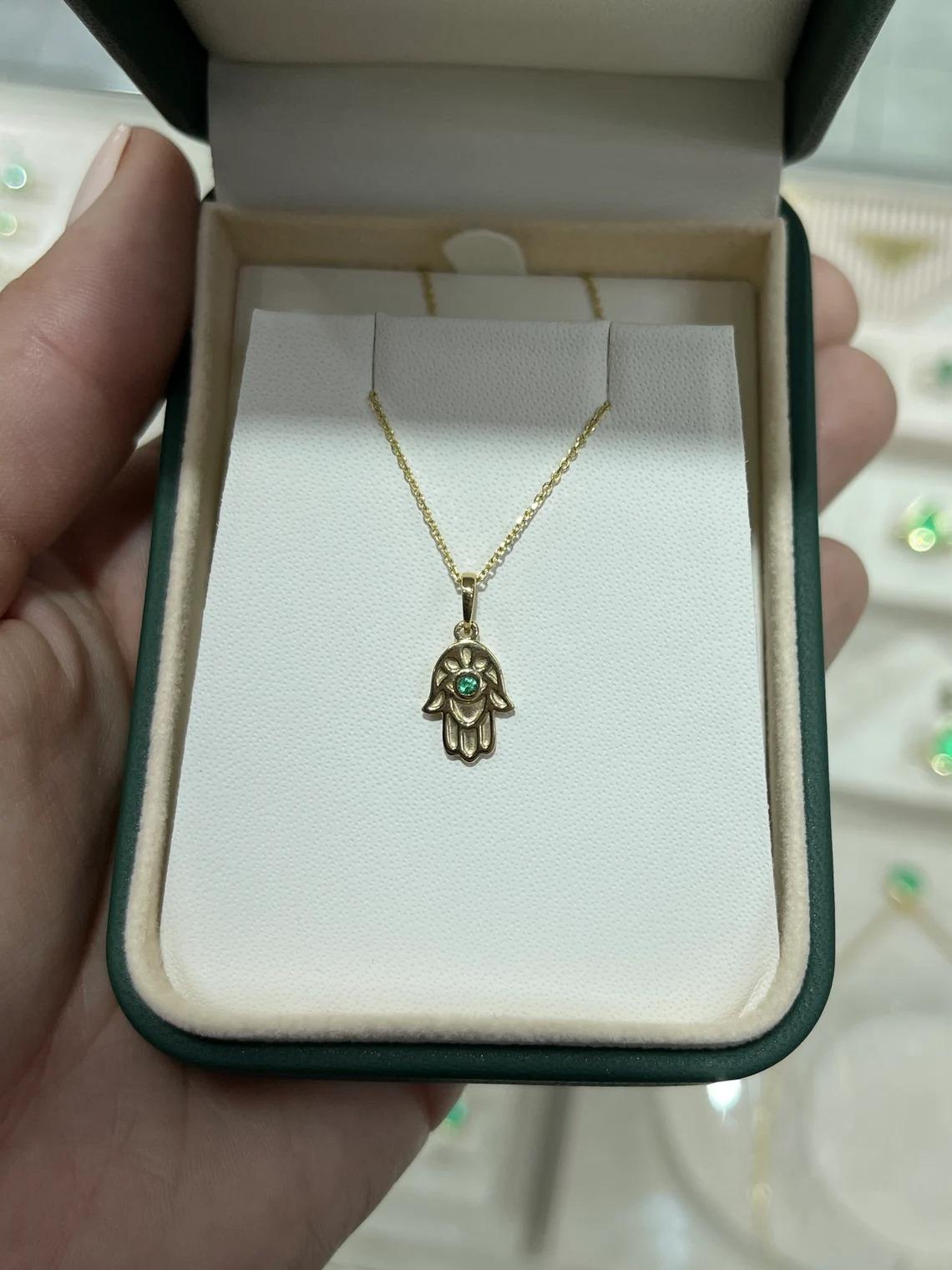 A gorgeous hamsa that features a stunning dainty round cut emerald in the very center. Skillfully crafted and set in 14K gold. The Hamsa (also known as Khamsa) is a hand-shaped amulet used for protection by both Jewish and Muslim people. Its name