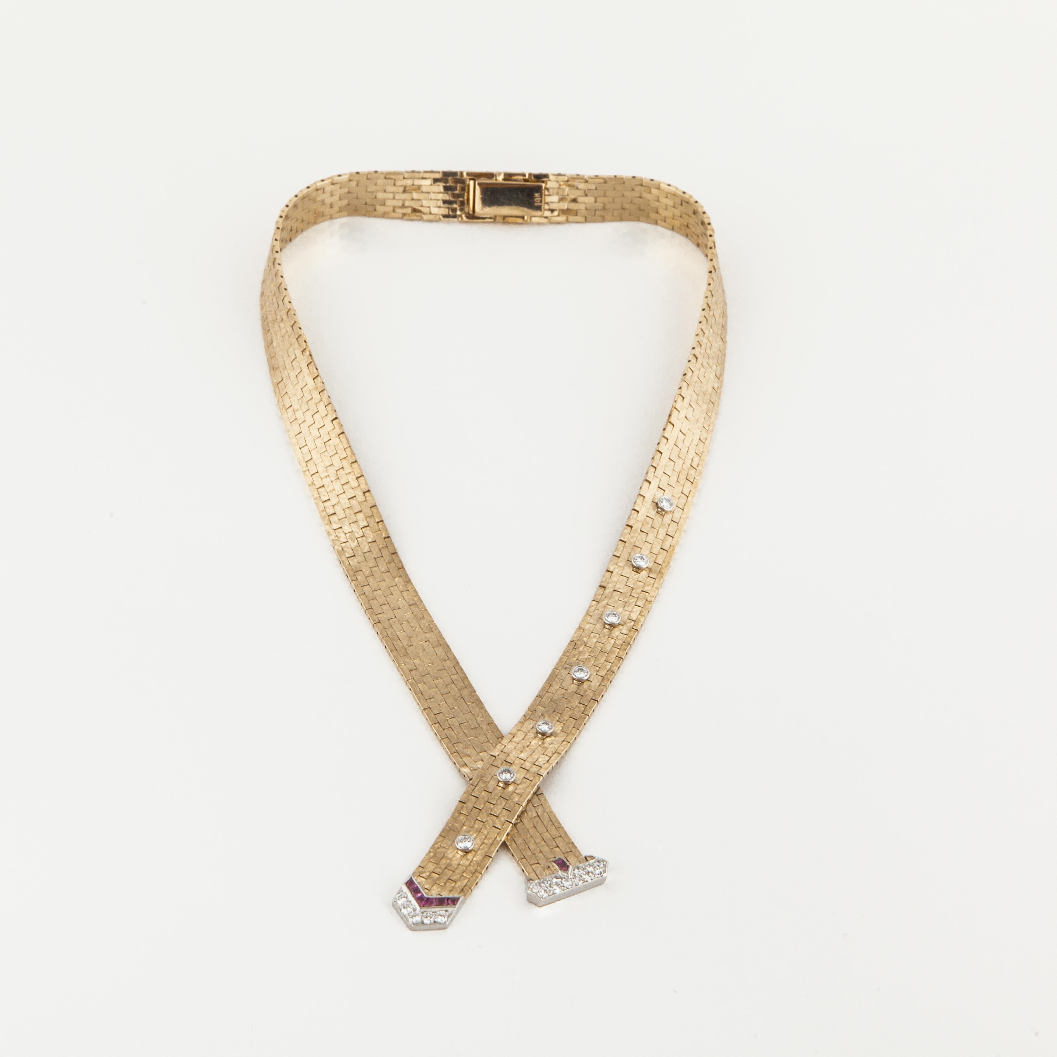 Retro buckle style necklace composed of 14K yellow gold with diamond and ruby accents.  There are two straps with a buckle at the ends, which criss cross at the bottom, and the top is brushed gold while the back is highly polished.  There are 24