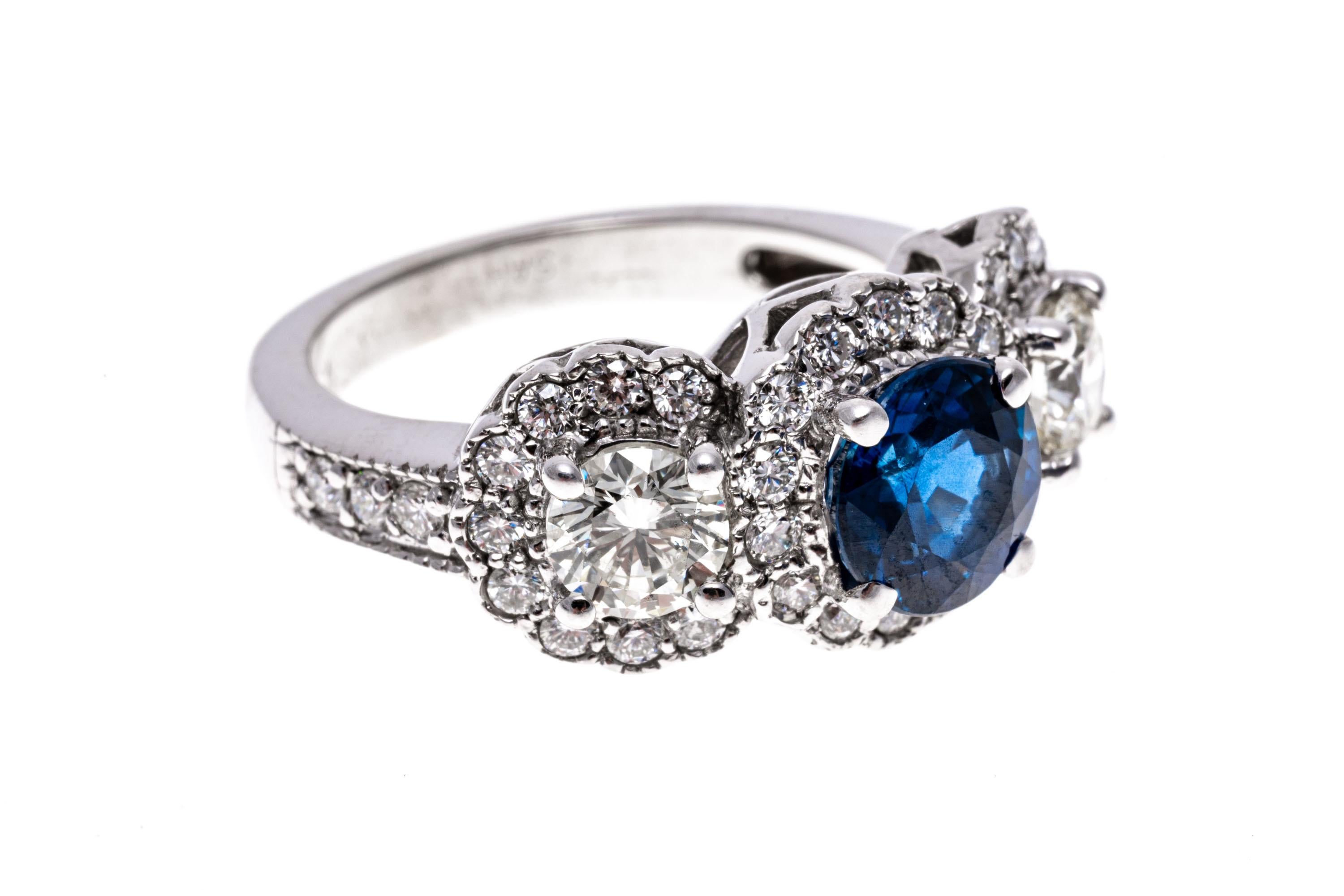 14k White Gold Sapphire And Diamond Halo Three Stone Ring.
This handsome ring is a three stone style, set in the center with a round faceted, medium blue color sapphire, approximately 1.44 CTS, flanked with a round brilliant cut diamond on each