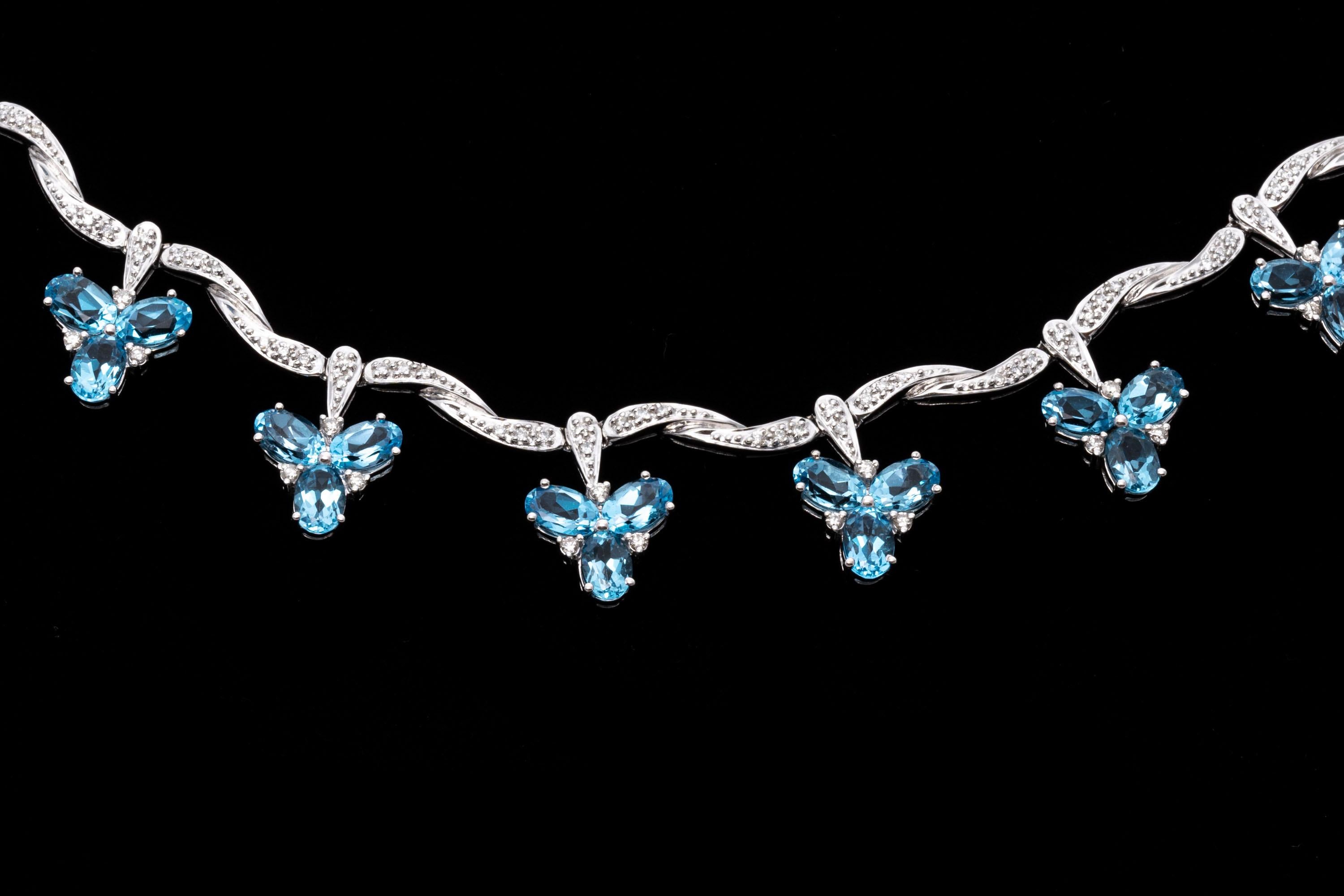 This interesting necklace is crafted of 14K white gold and presents an elegant design. Twist style links with diamonds support small charms set at intervals over the front of the necklace. These charms are set with small clusters of oval cut topaz