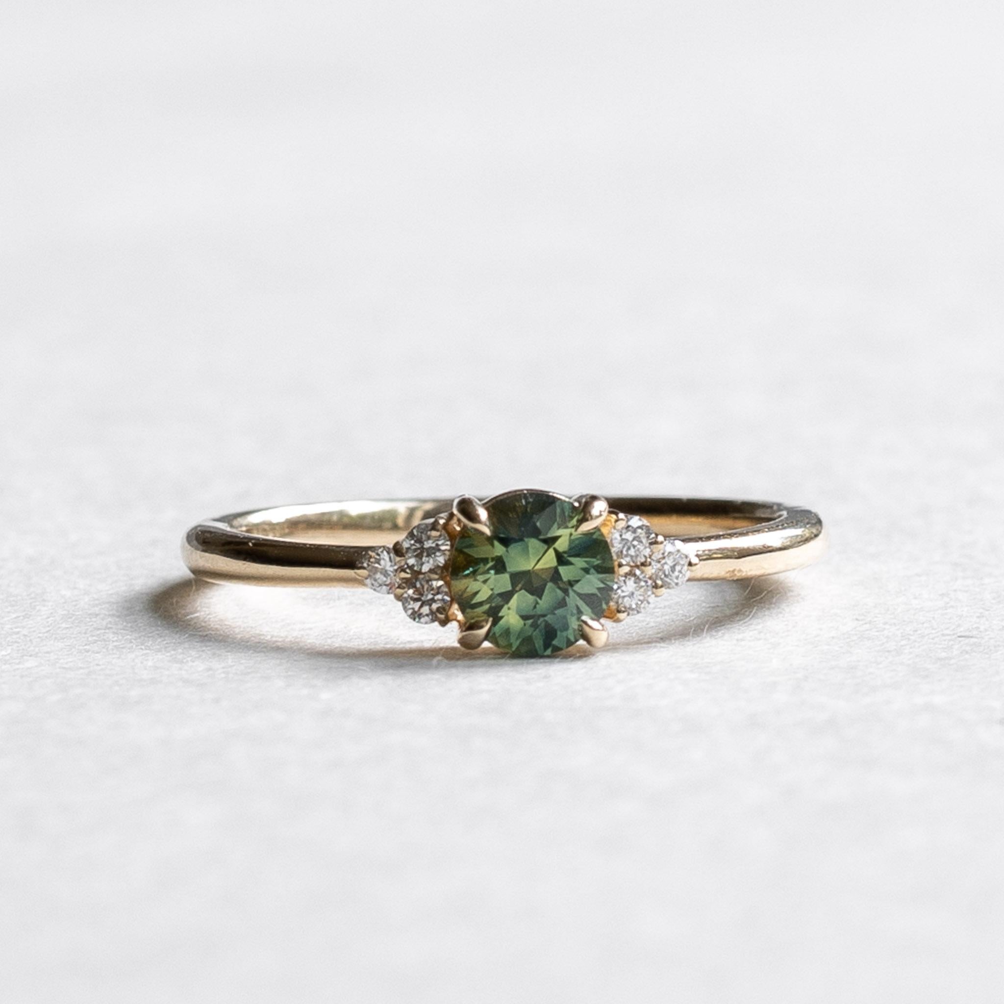 Australian teal sapphire with diamond accents 
Stone: Parti Sapphire
Metal: 14k Gold
Stone Shape: Round
Stone Size: 5mm
Stone Weight: 0.589 carat
Accent Stone: Natural Diamond
Accent Color and Clarity: G+, VS
Total Diamond Weight: 0.12 carat
Band