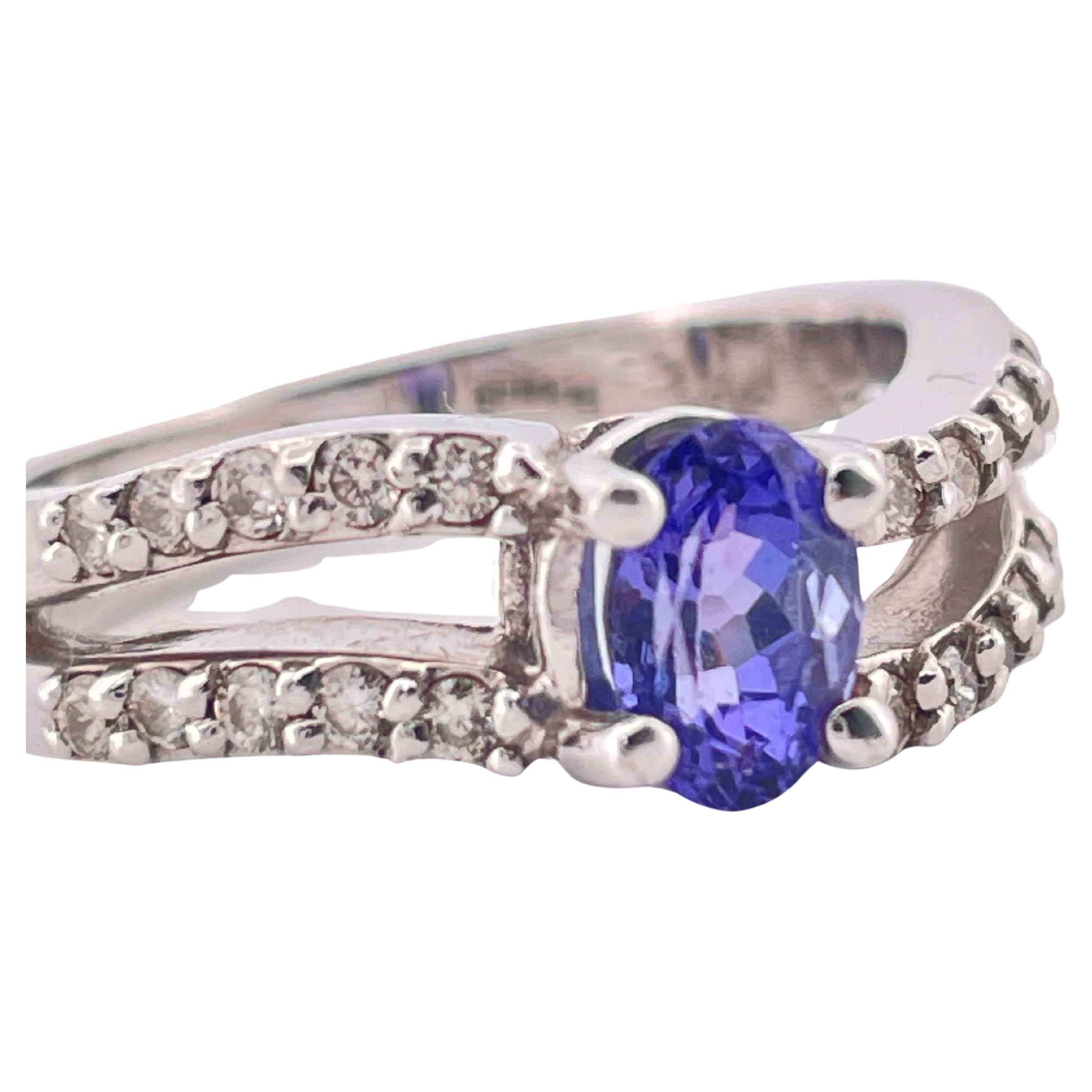 Este anillo de oro blanco de 14 quilates, que pesa 5,4 gramos, exhibe una impresionante tanzanita talla oval de 0,60 quilates, apreciada por su intenso tono azul violáceo y su excepcional claridad. La piedra central está elegantemente sujeta por
