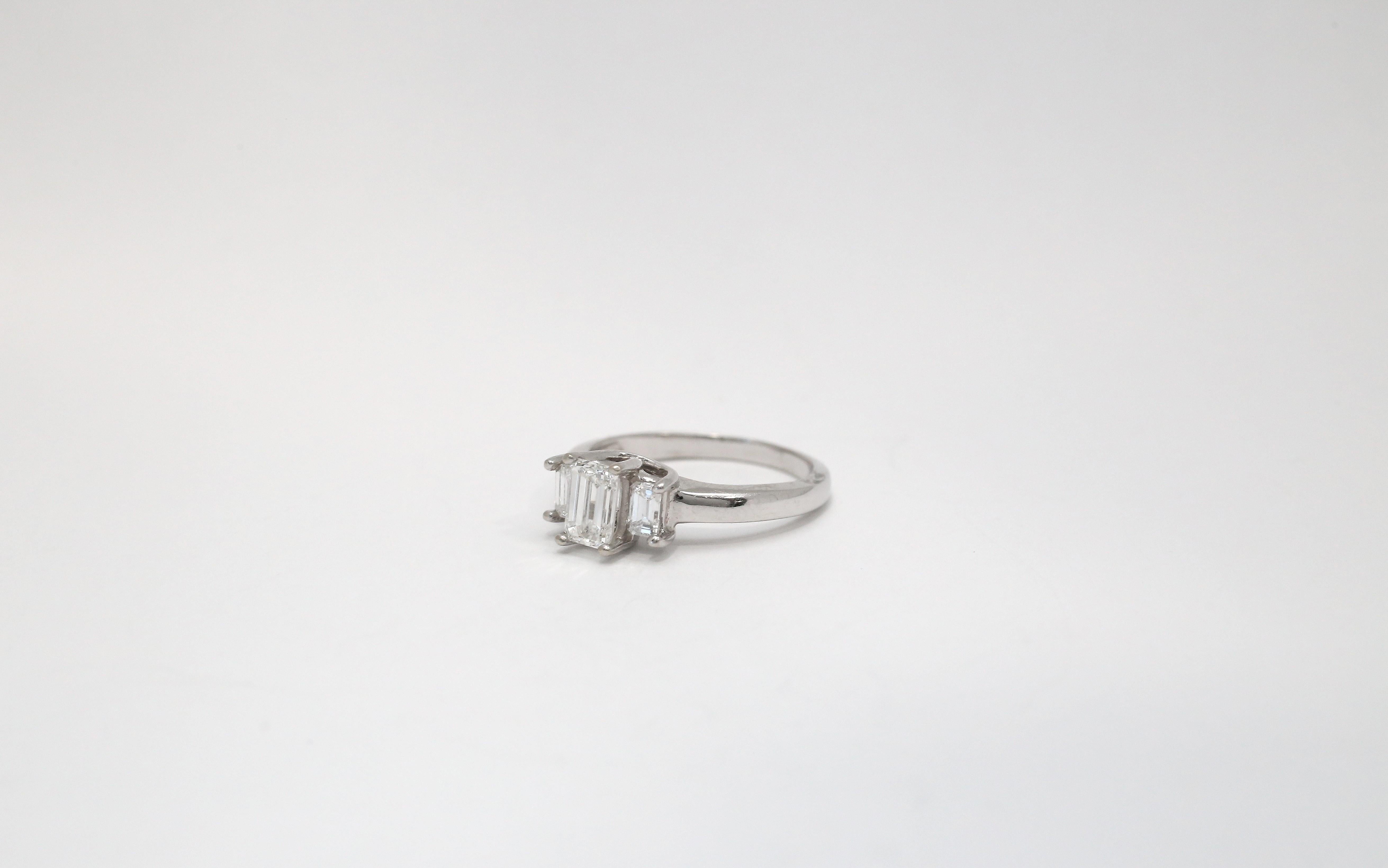 This elegant 14k white gold engagement ring showcases a timeless three-stone design set in a graceful trellis setting. At the center is a striking emerald-cut diamond, flanked by two matching emerald-cut diamonds that enhance the ring’s