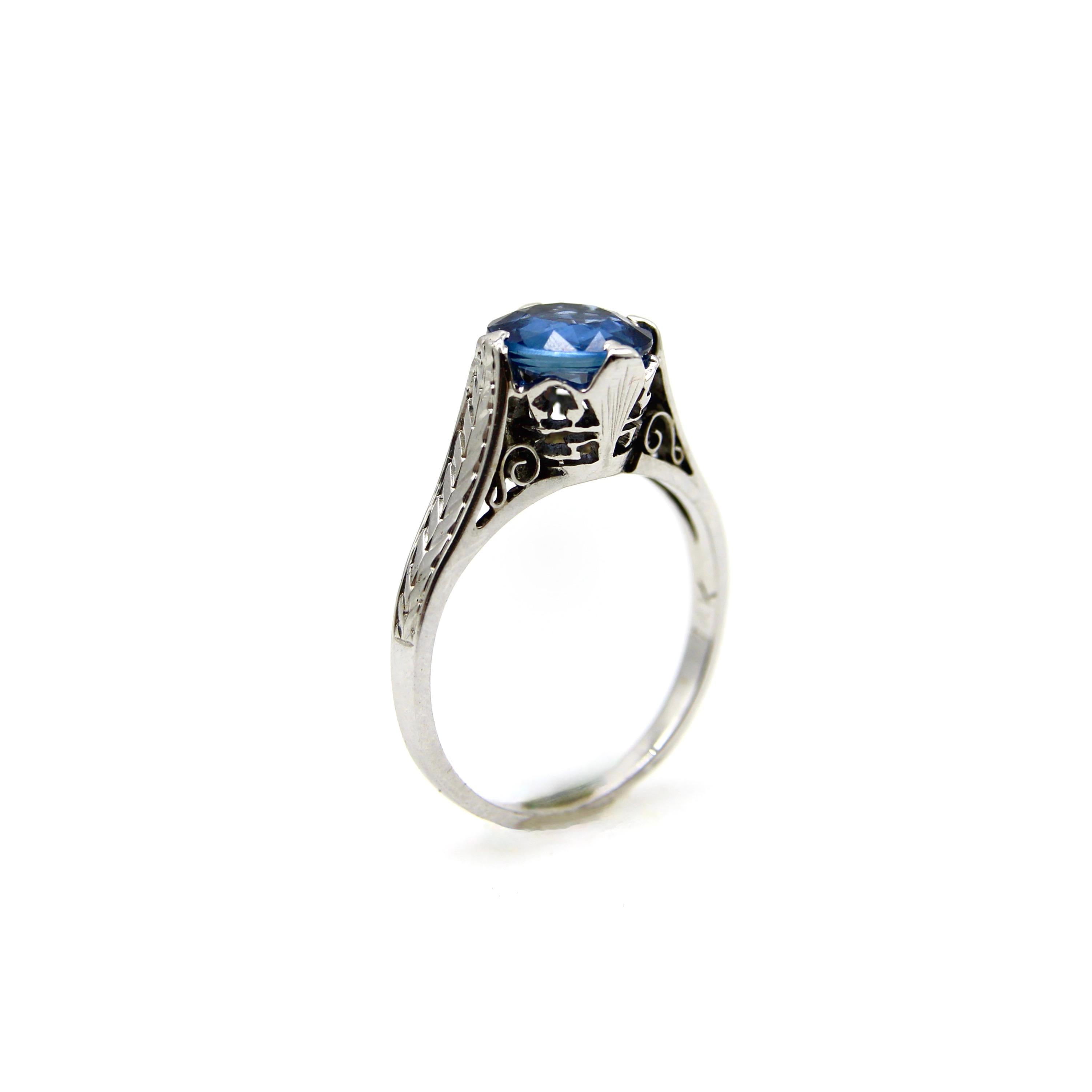 This 14k white gold ring features a natural blue sapphire in an Art Deco mount. The sapphire is a gorgeous shade of medium blue with grayish undertones. In the sunlight, the stone sparkles with an even more vibrant shade of blue, and the stone’s cut
