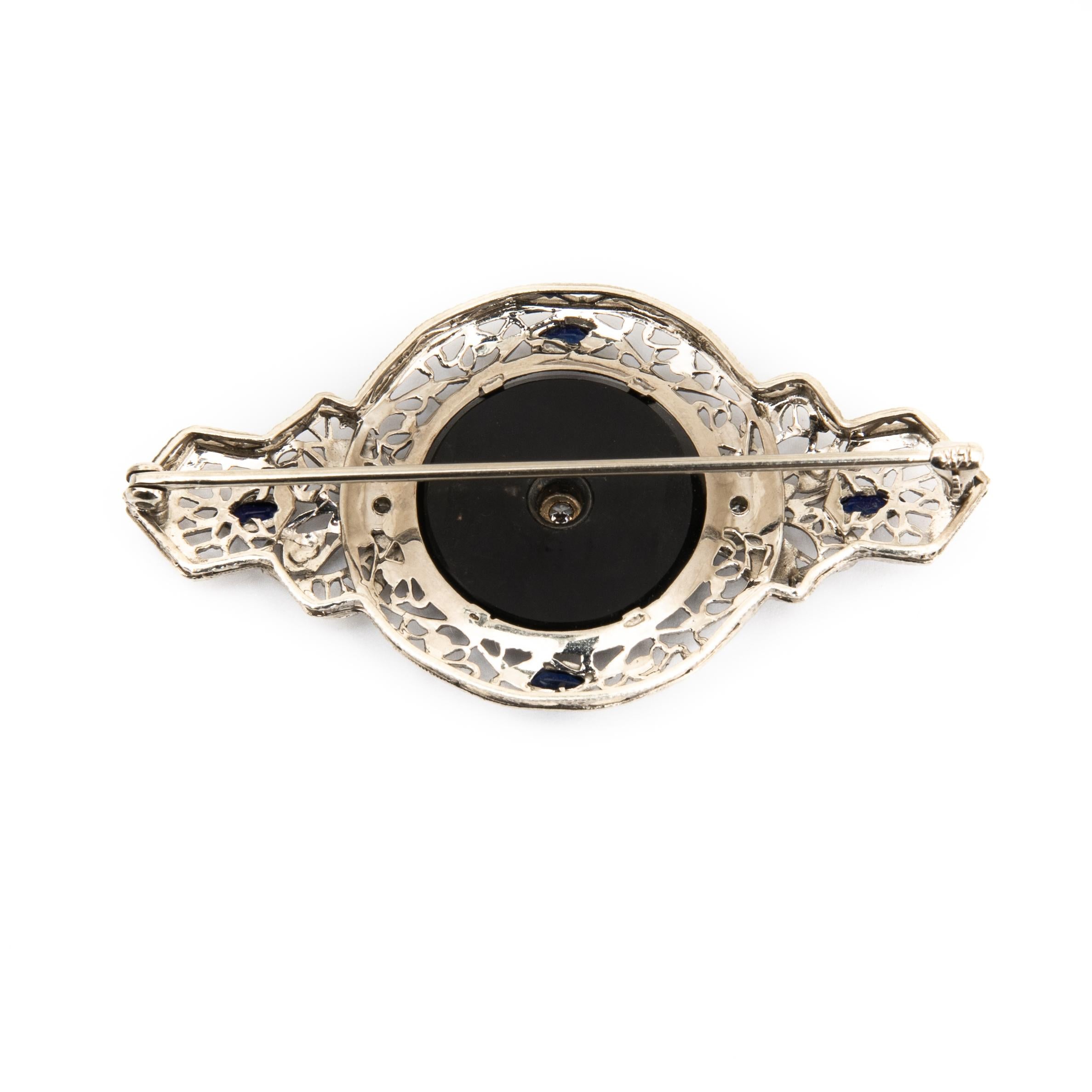 Offered is An Art Deco 14K white gold black onyx, diamond and synthetic blue sapphire brooch. This brooch features a black onyx round tablet, which has an old European cut diamond in the center and a diamond accent on each side. The border of the