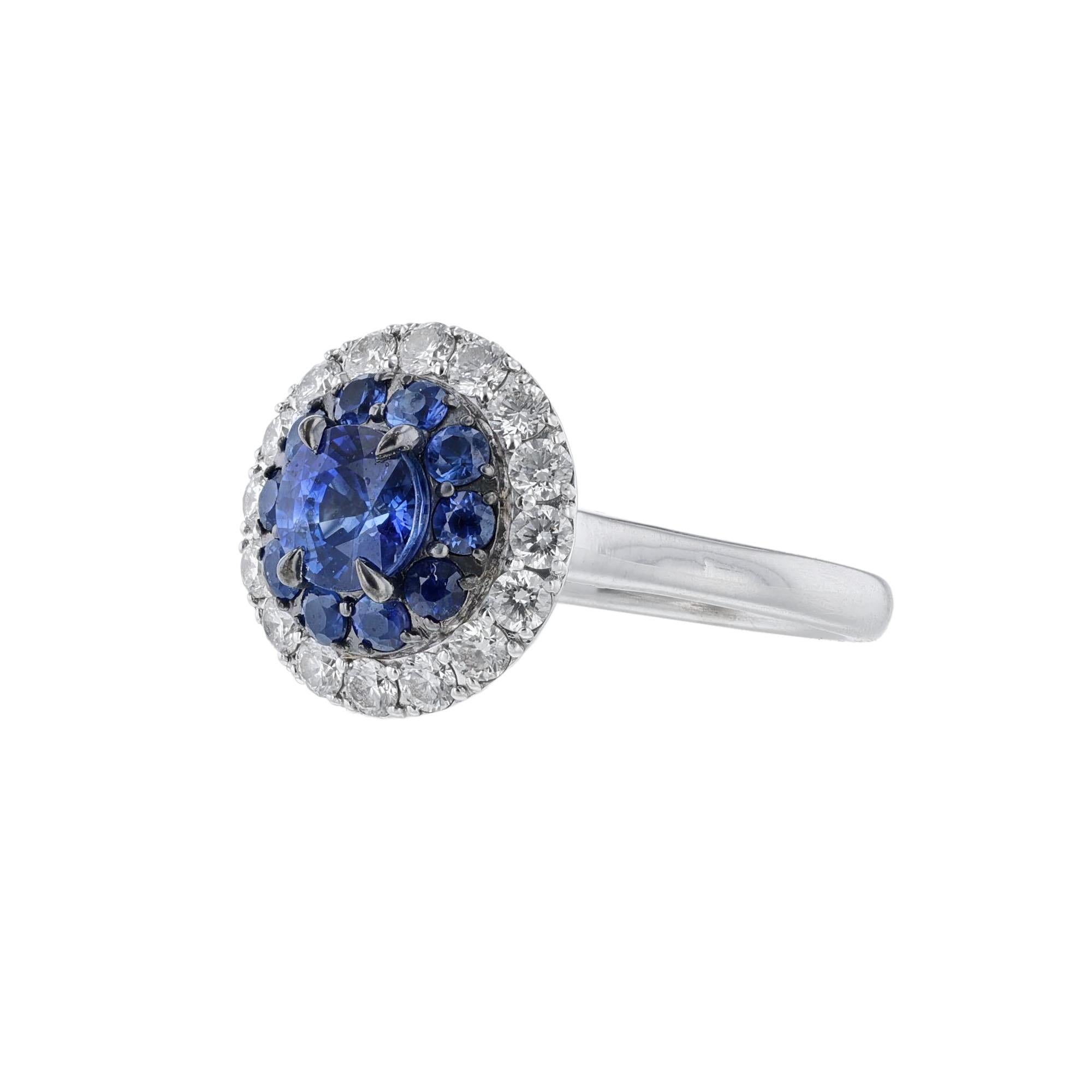 This ring is made in 14K white gold and features 1 center round blue sapphire weighing 1.12 carats. Surrounded by a halo of round cut blue sapphires weighing 0.55 carat. A secondary halo of round cut diamonds and 10 diamonds on each side of the