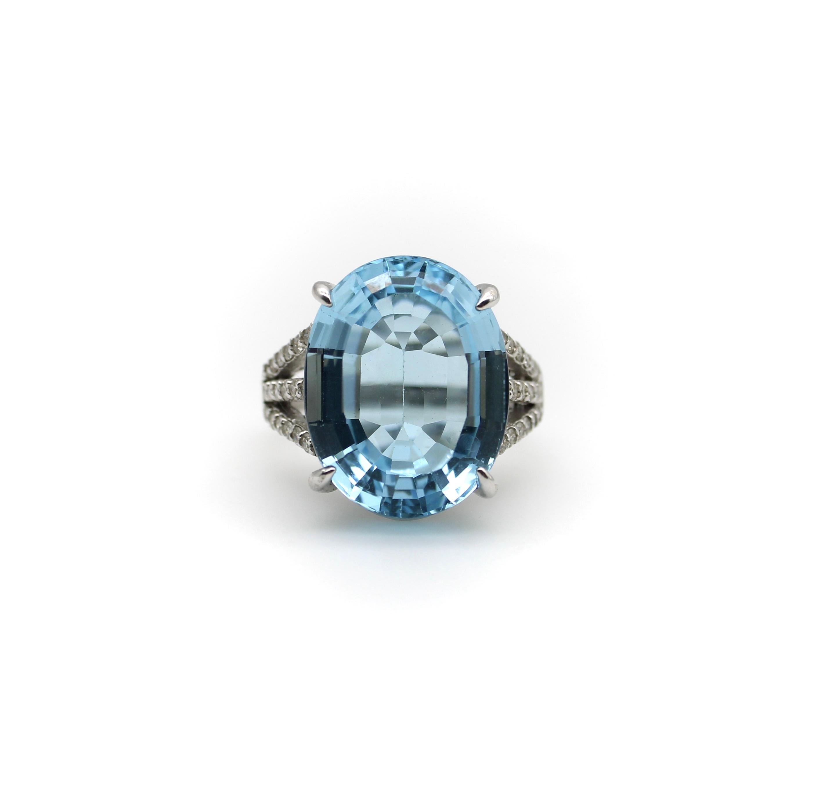 This 14k white gold, diamond, and blue topaz ring contains a striking 19+ carat vibrant blue gemstone. The oval stone is beautifully cut with facets that reflect a myriad of Mediterranean blue magic. An elaborate, scrolling foliate pattern details