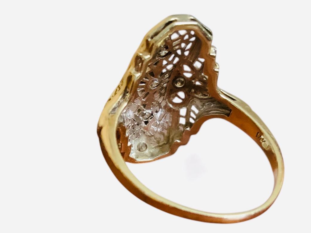 This a 14K Gold Diamond Art Deco Ring. It depicts a white gold rectangle made of filigree and decorated with five diamonds in pave setting in a column. Each of the diamonds has a diamond shaped frame. A quatrefoile adorns the side of the ring and