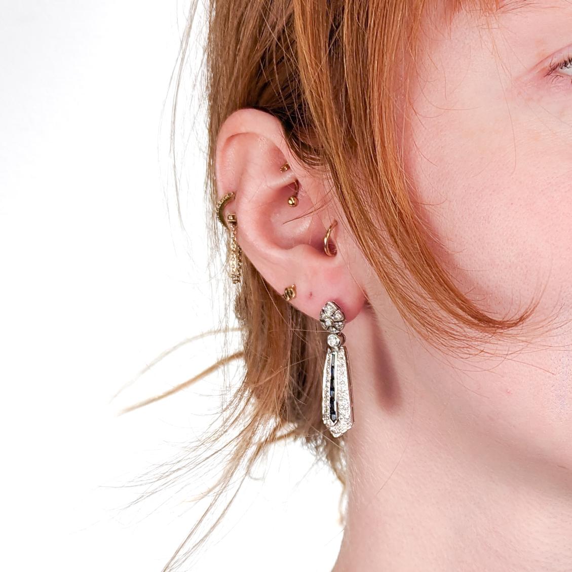 A fine pair of Art Deco style earrings.

In 14 karat white gold with white diamonds & synthetic blue sapphires.

Each earring with a pear shaped flora-form top supporting pendant drops. 

The tops are set each with 7 white diamonds, and the drops