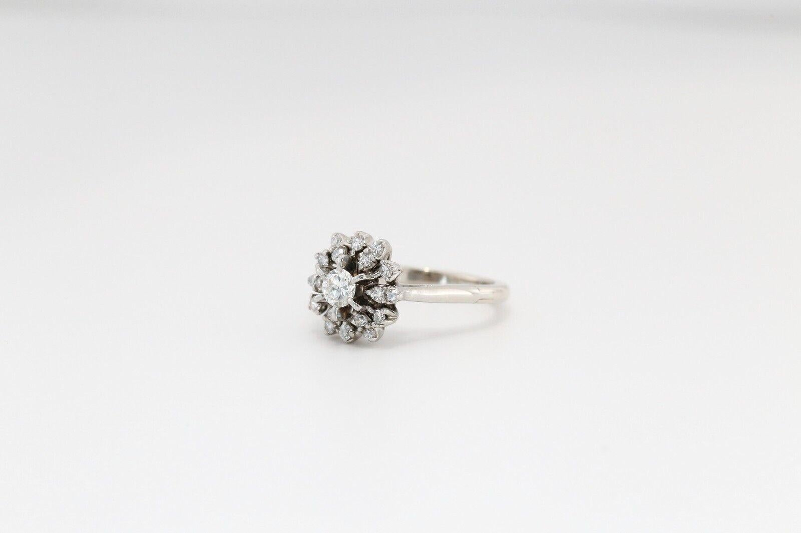 Cette élégante bague à diamant en or blanc 14k associe une brillance classique à une ingénierie intelligente. Doté d'un éblouissant diamant rond central entouré d'une grappe florale de pierres d'accent étincelantes, le design évoque une