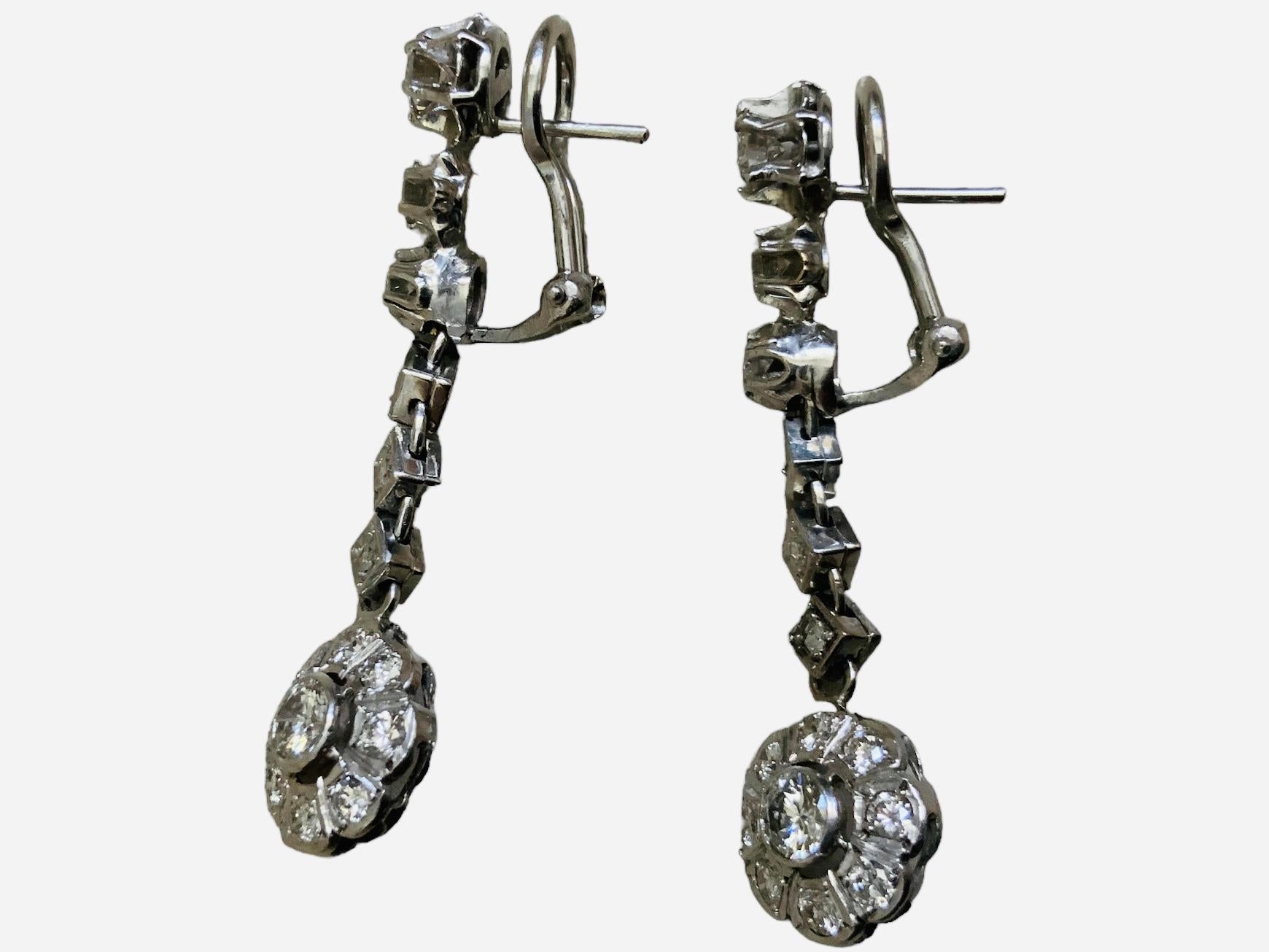 This a Pair of 14K White Gold Diamonds Earrings. It depicts a pair of dangle earrings consisting of a chain made of graduated round diamonds mounted in prong and pave settings ending in a flower with a large round diamond on bezel setting in the