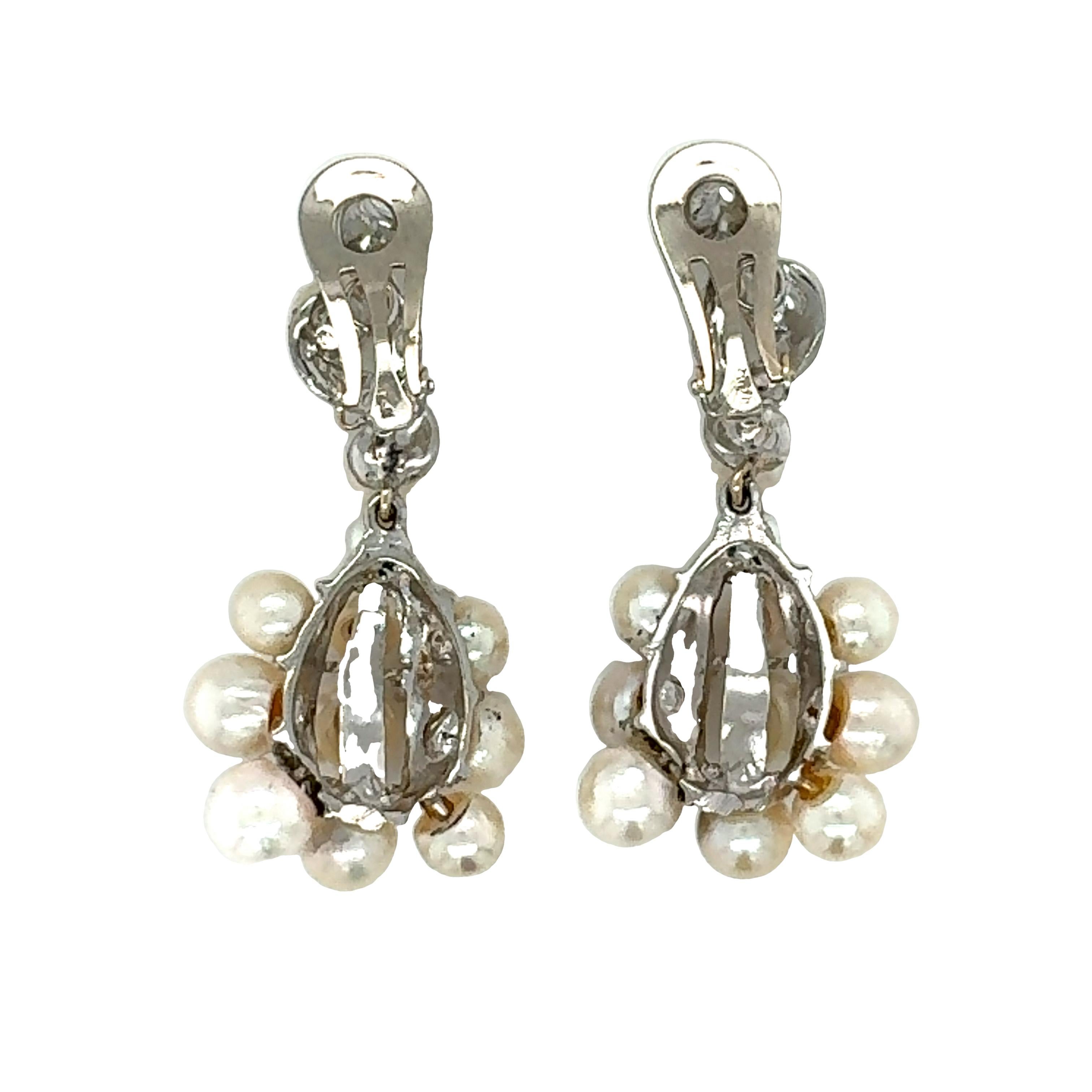 One pair of pearl and diamond dangling earrings in 14K white gold featuring 24 white, round cultured pearls measuring 4.00-6.50 millimeters in diameter. Accented by 8 prong set, single round cut diamonds totaling 0.12 ct. with L-M color and SI-2,