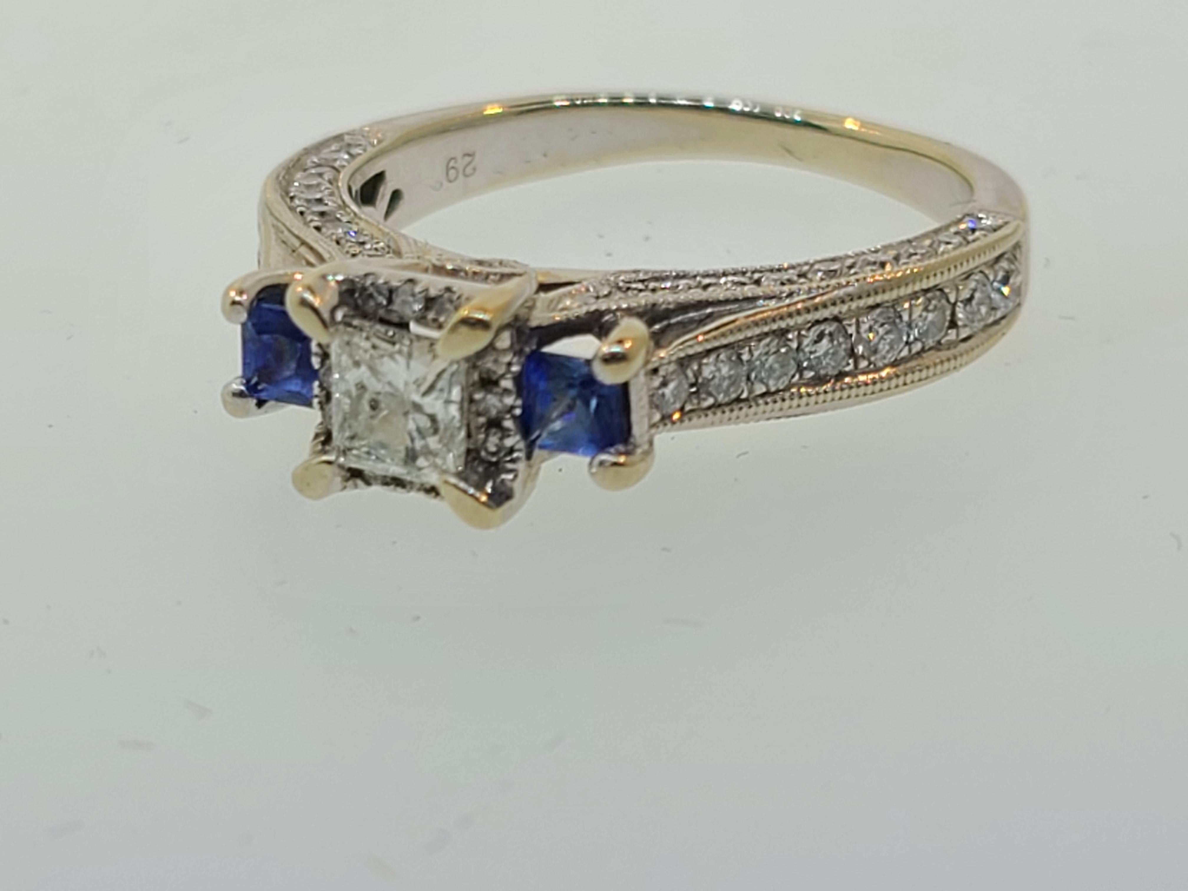 This 14k white gold engagement ring features a  4.20mm princess cut diamond center solitaire secured in a four-prong setting complemented by a recessed halo of nine round brilliant cut diamonds for added dimension. Two square cut blue sapphires give